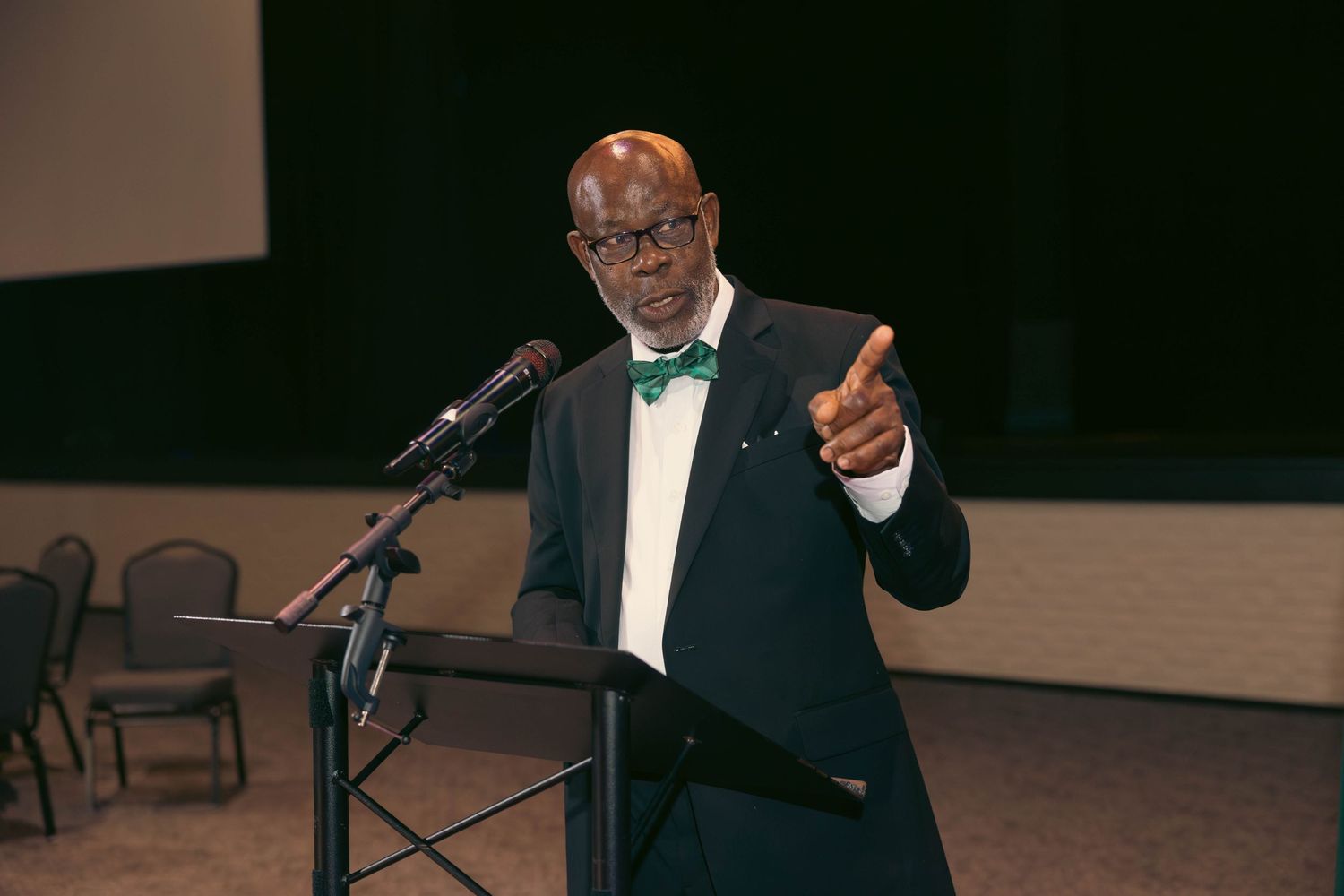 Elderly man in tuxedo with green bow tie points while speaking at podium.