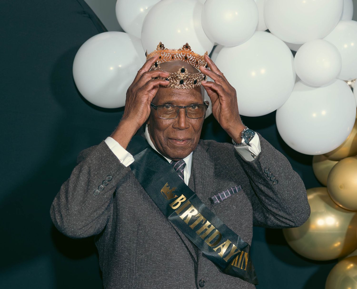 Elderly man wearing birthday sash places decorative crown on his head at party.