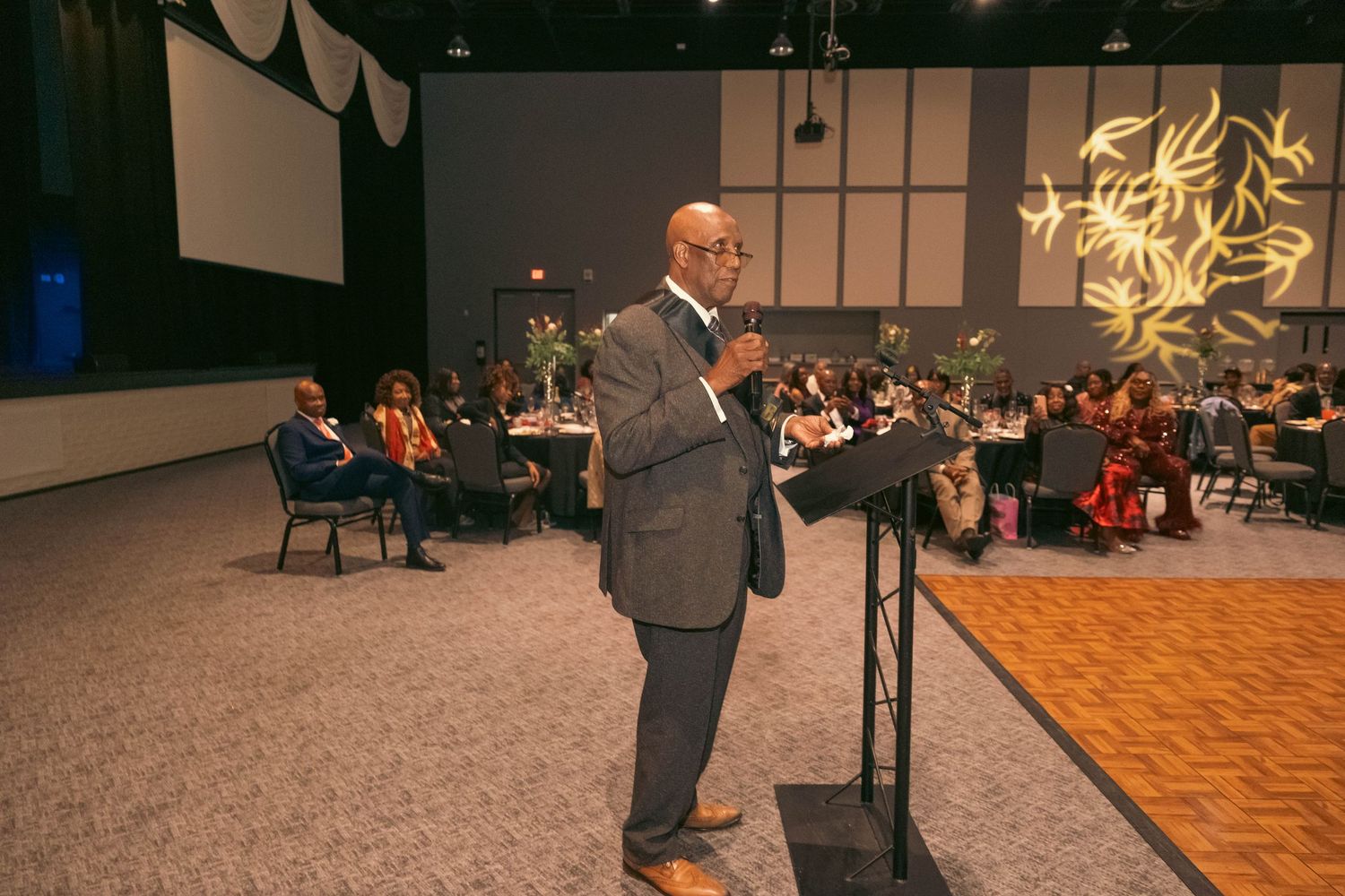 Man in gray suit speaking at podium before seated audience in decorated event hall.