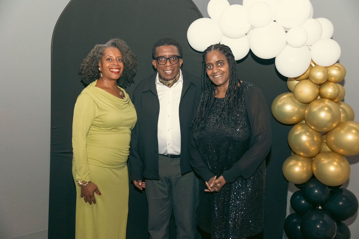 Three people posing together in front of balloon arch at formal celebration event.