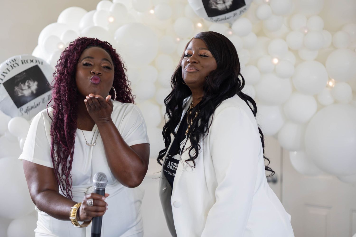 Two women posing together at a baby shower event with white balloon arch backdrop, one holding microphone.