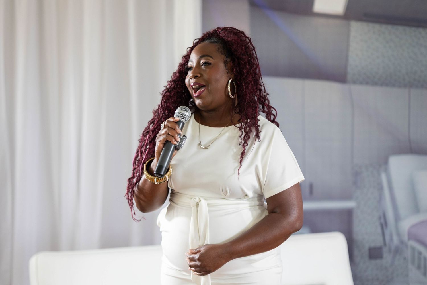 A woman in a white dress speaks into a microphone at an indoor event.