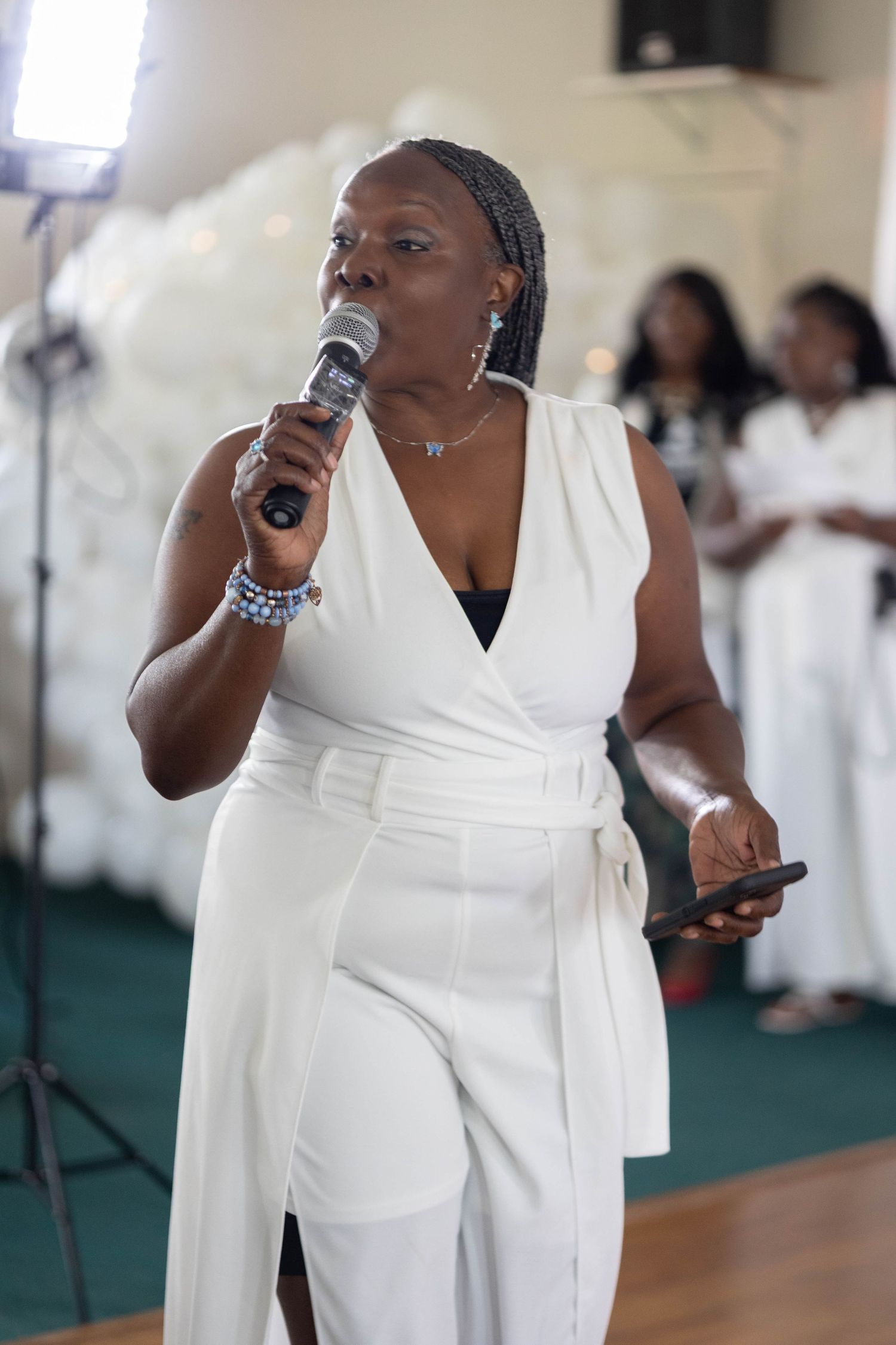 A woman in a white jumpsuit speaks into a microphone while holding a phone at an event.