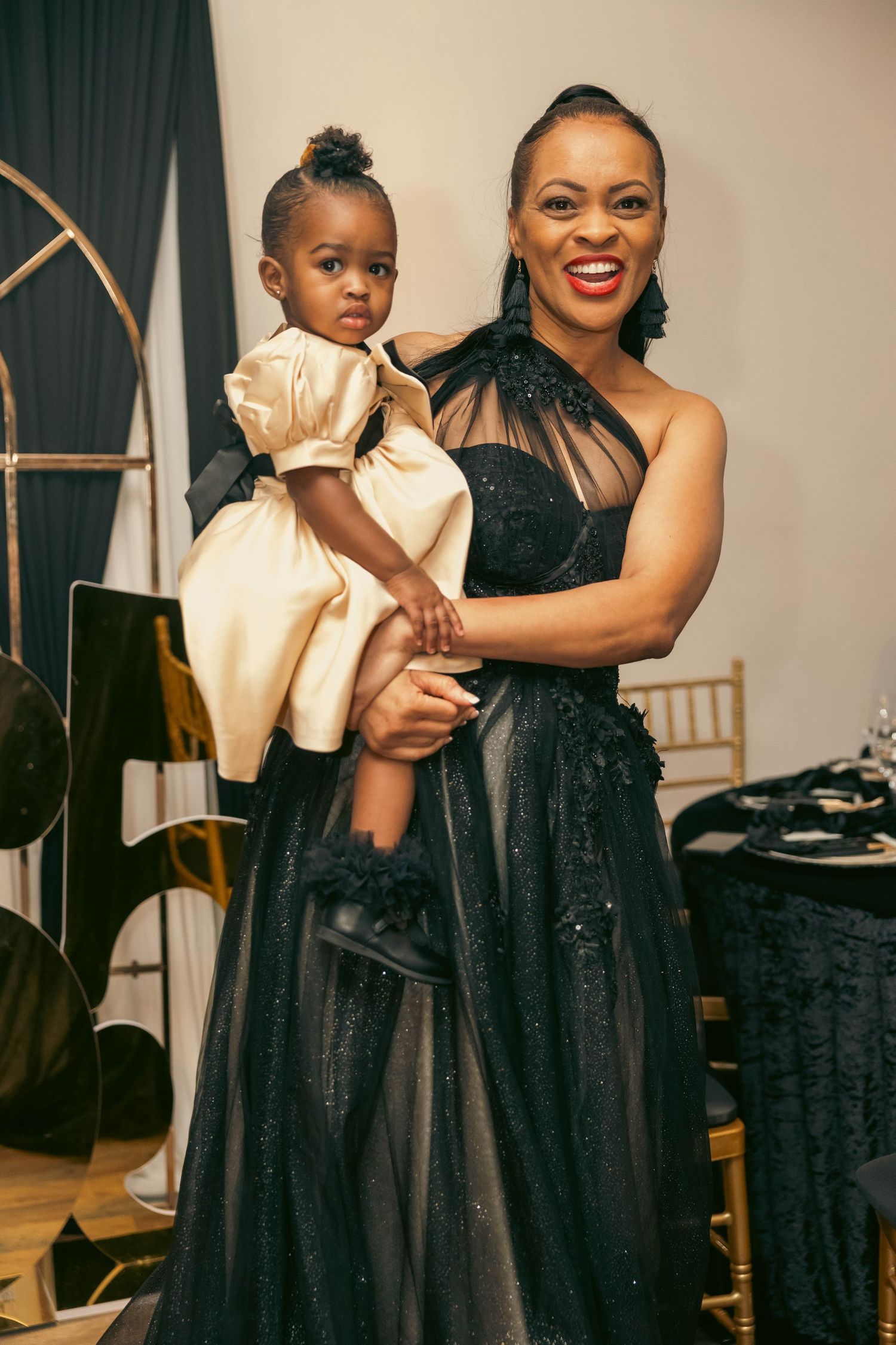 Smiling woman in black gown holding a young girl in cream dress at an elegant formal celebration.