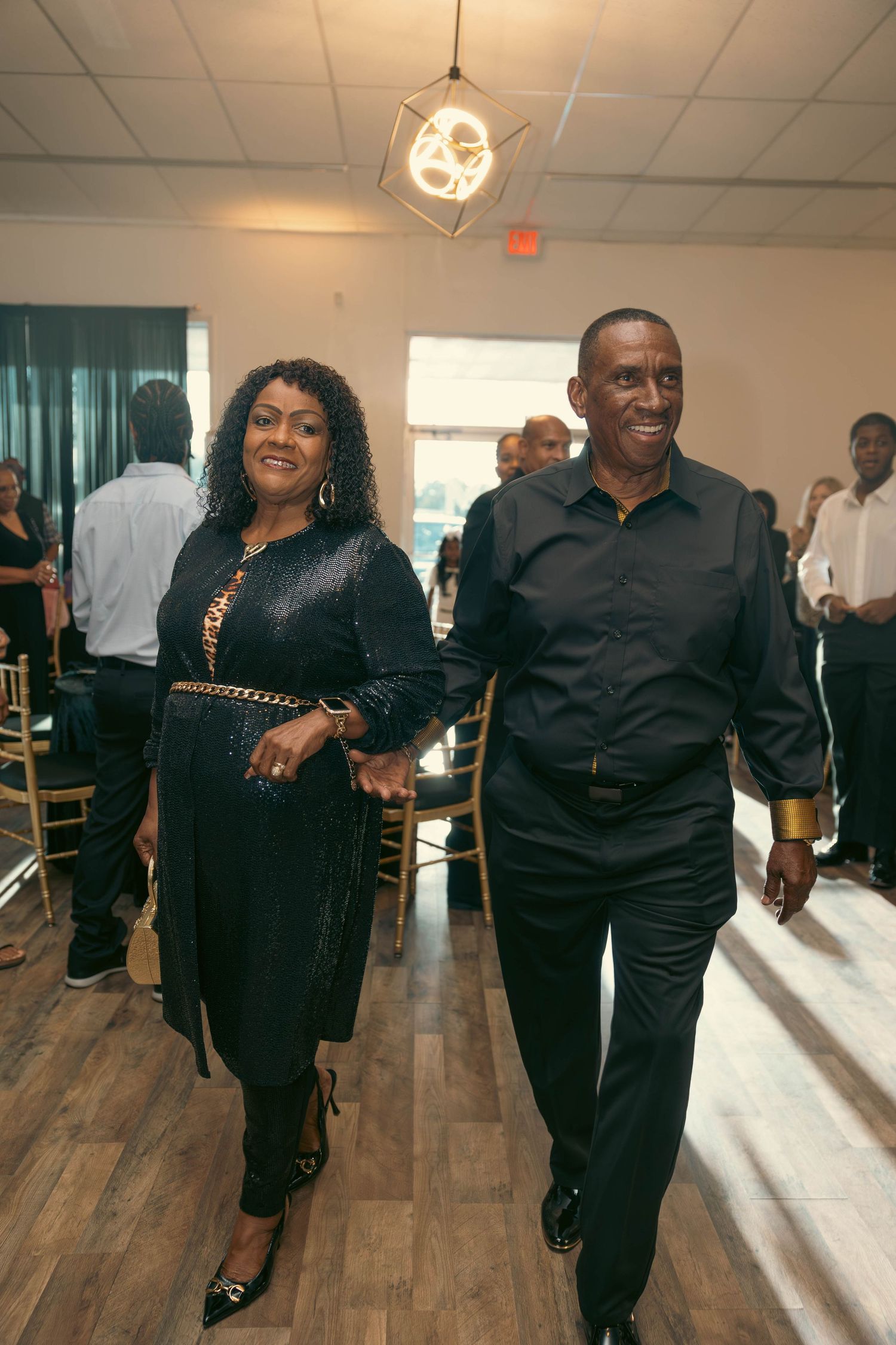 Elegantly dressed couple in all-black attire walking hand in hand at a formal celebration event.