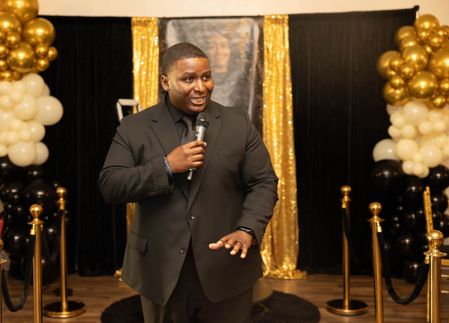 Man in black suit speaking into microphone at black and gold balloon-decorated formal event venue.