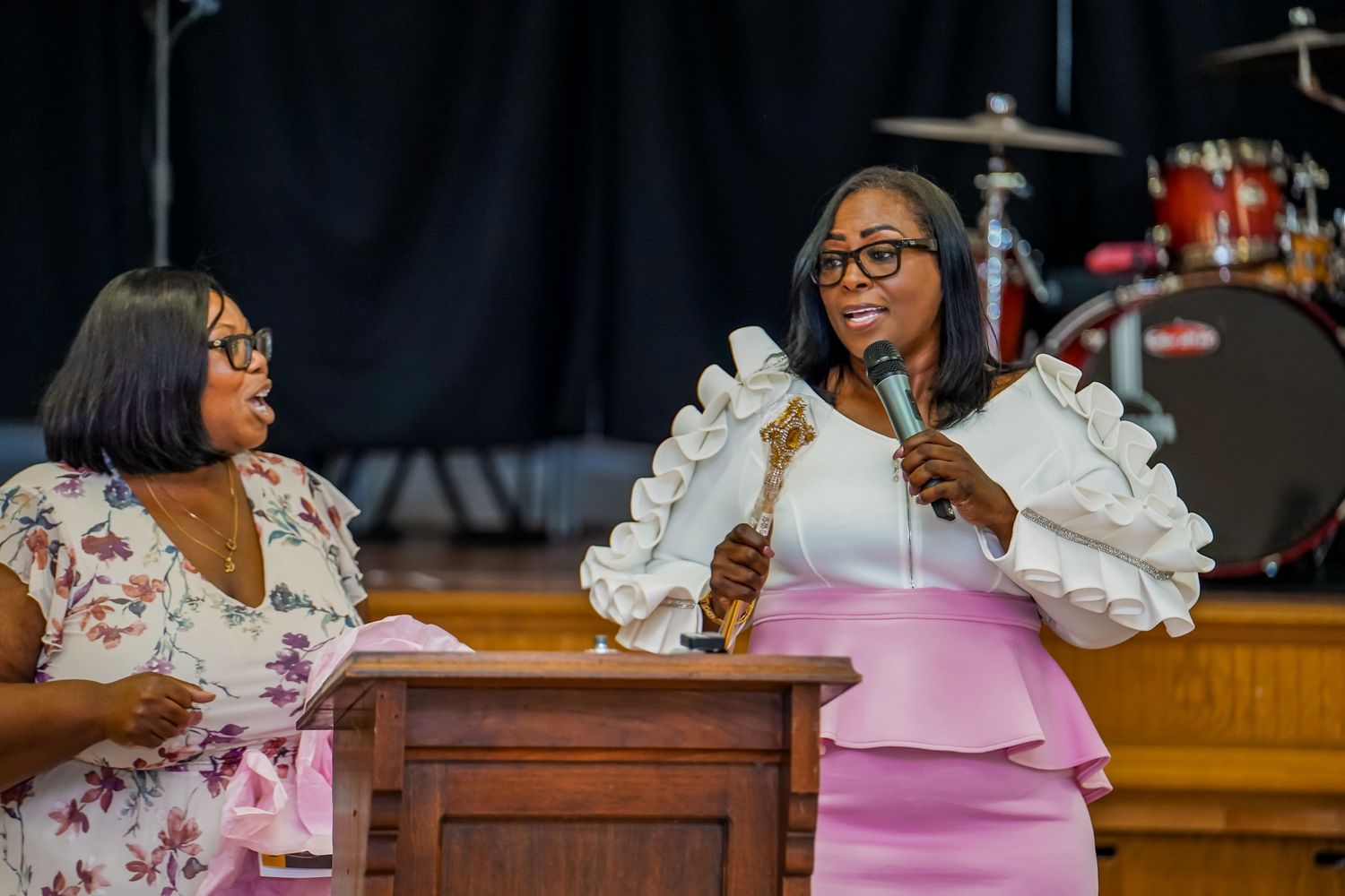 Woman in white peplum dress speaks at podium holding microphone during church ceremony on stage.