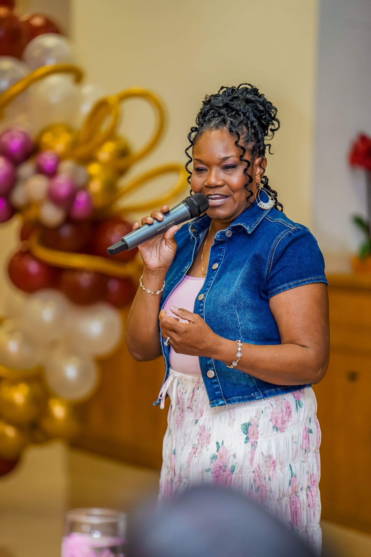 Woman in denim jacket sings into microphone at festive event with balloon decorations behind her.