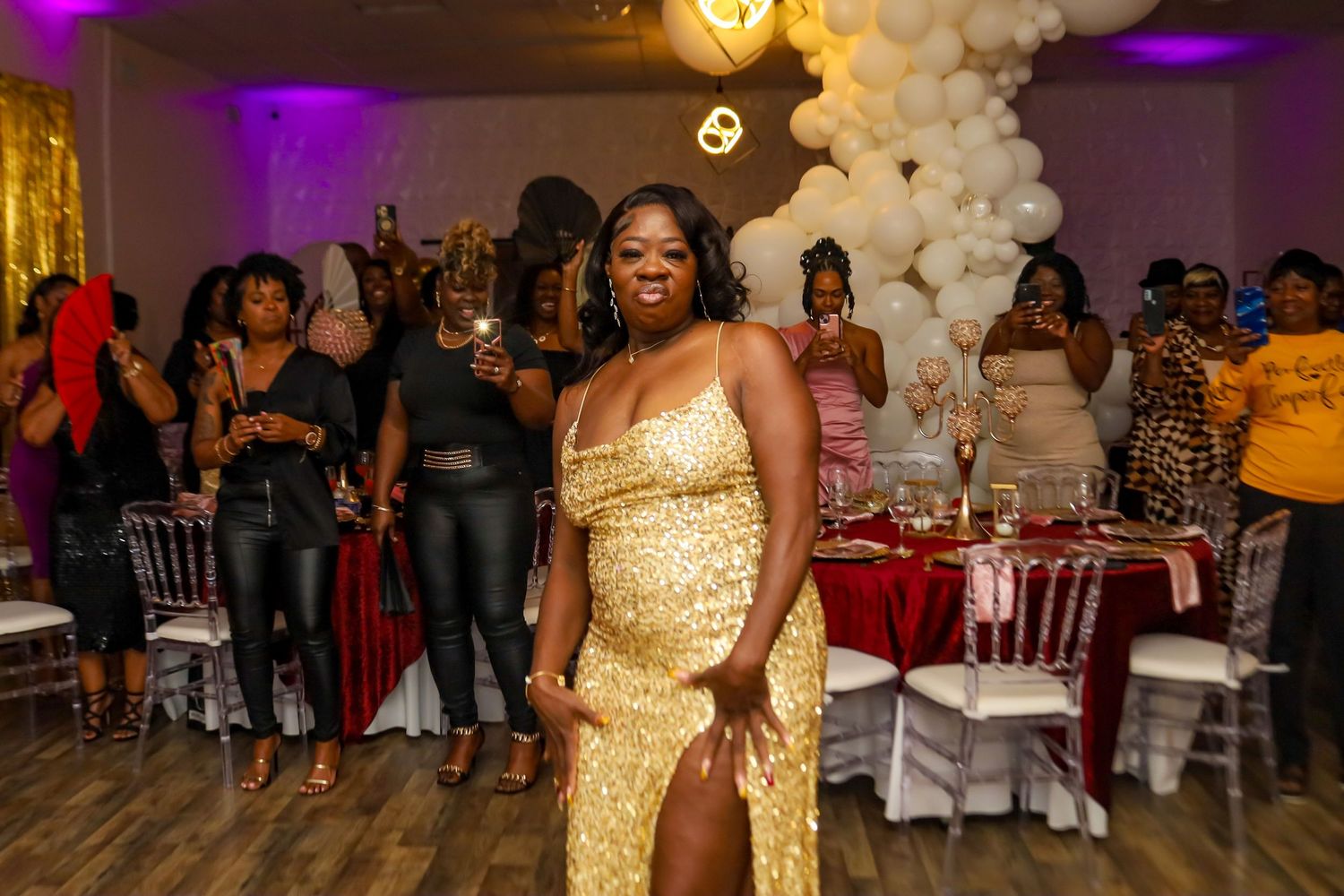 Woman in gold sequin gown posing at a festive celebration with red and white balloon decor.