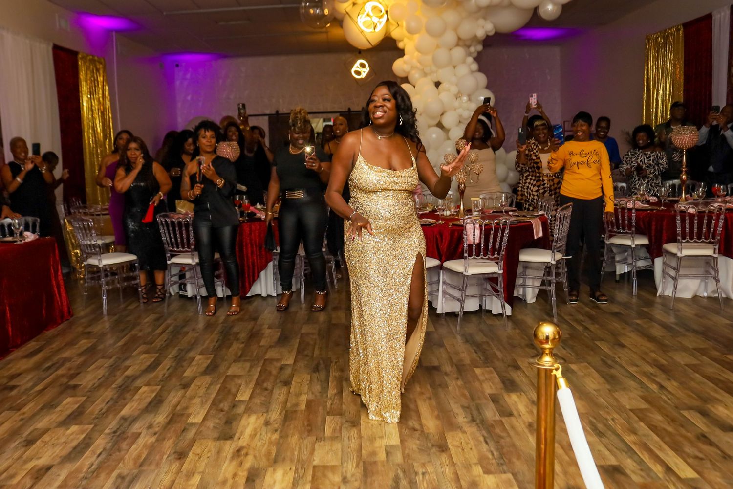 Woman in gold sequin gown walking through a decorated event hall with white balloon arch backdrop.