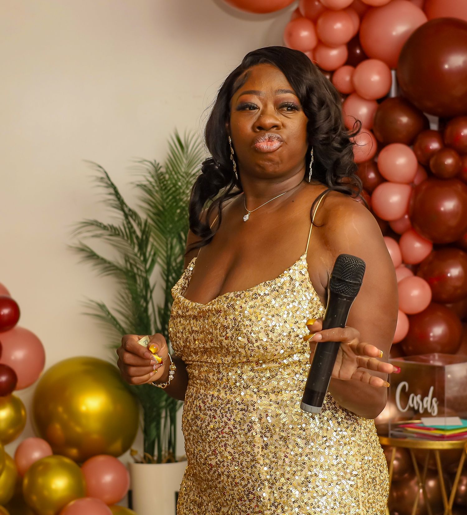 Woman in gold sequin dress holding a microphone at a celebration with balloon garland backdrop.