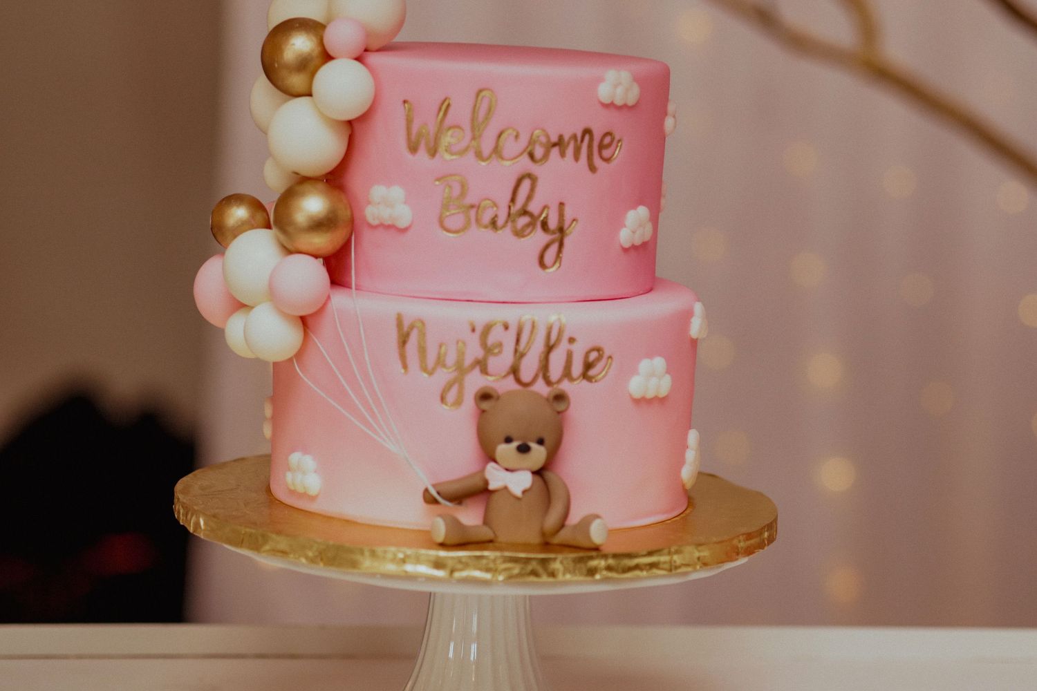 Two-tier pink Welcome Baby NyEllie cake with teddy bear and balloon decorations.