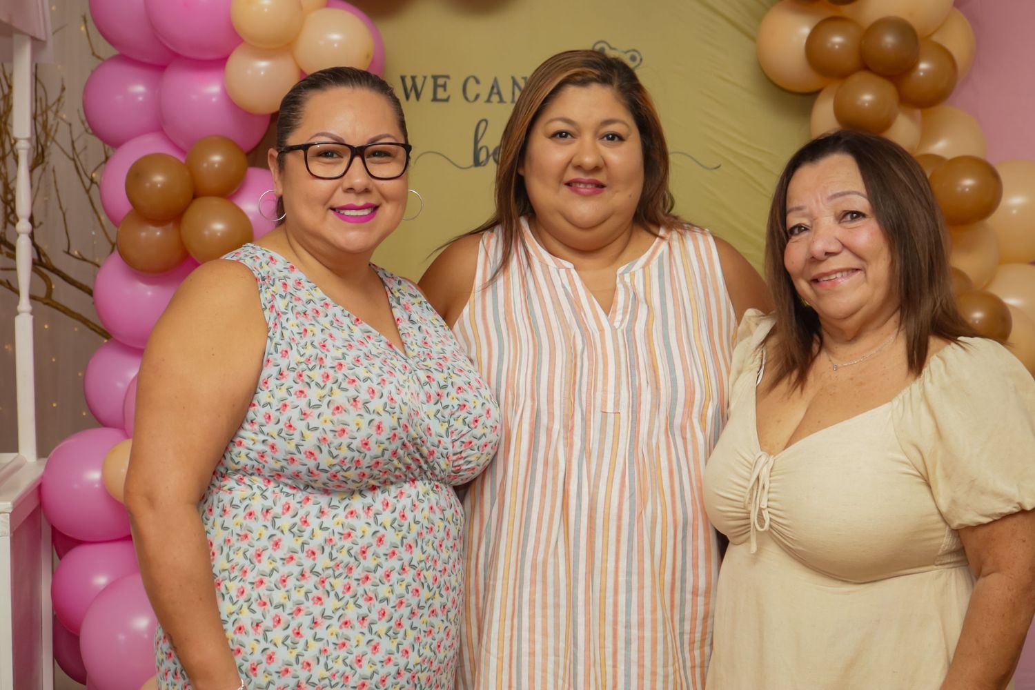 Three women smiling together in front of pink and gold balloon arch at baby shower.
