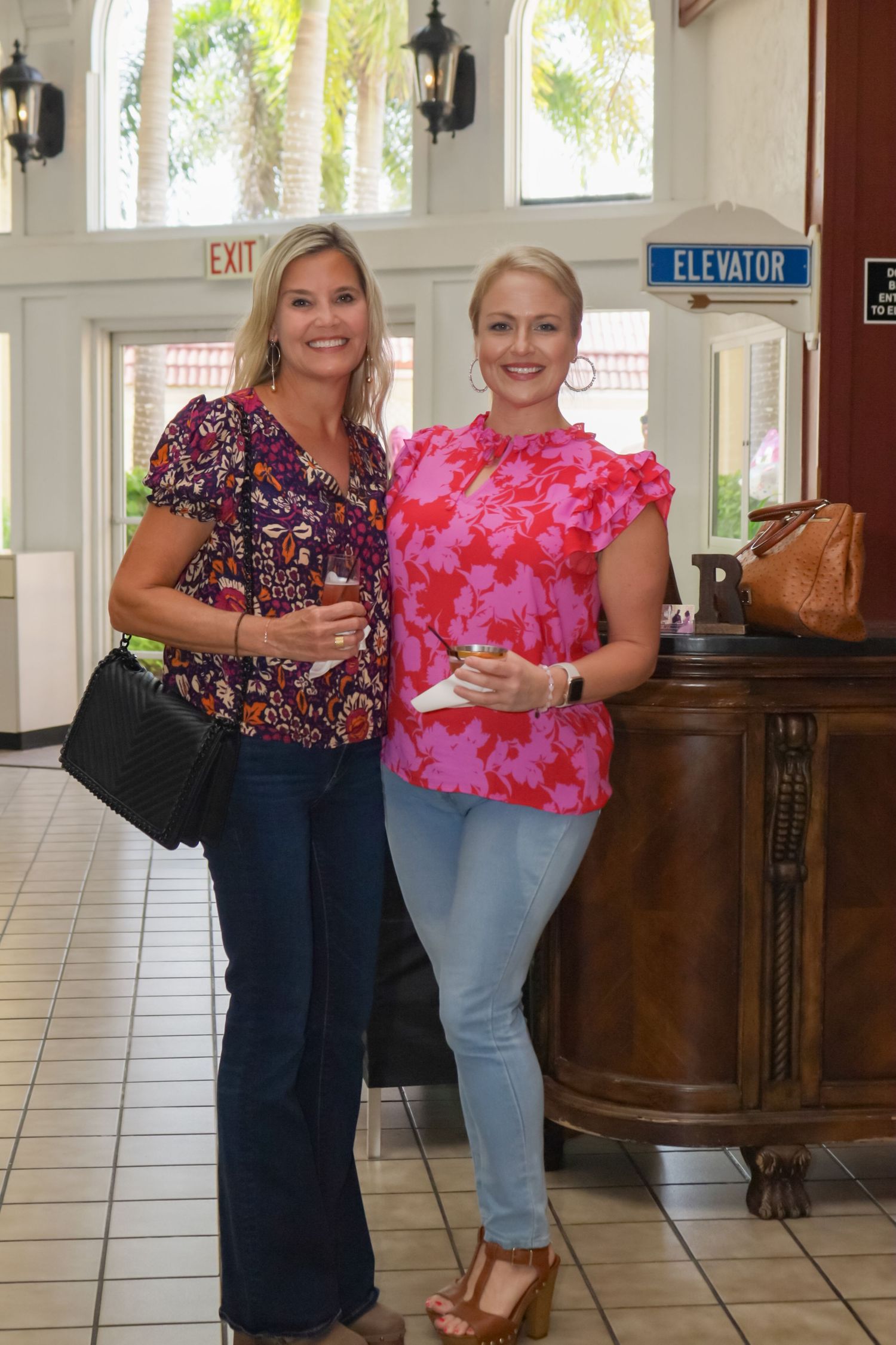 Two women holding drinks and smiling in floral tops at indoor venue with elevator sign.
