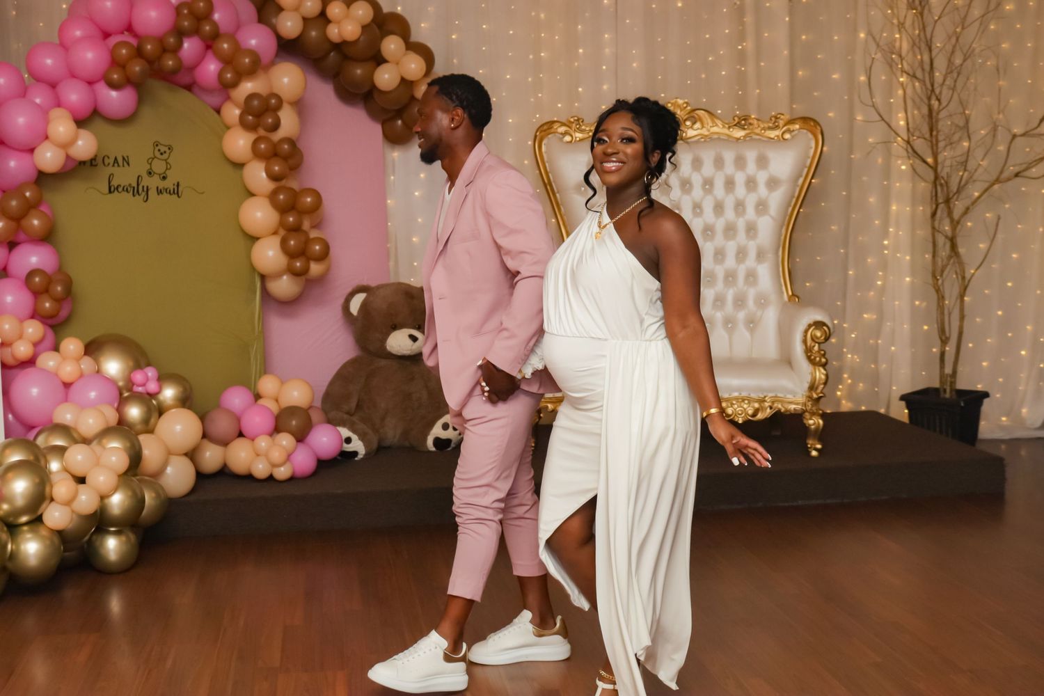 Pregnant woman in white dress dances with man in pink suit at baby shower.