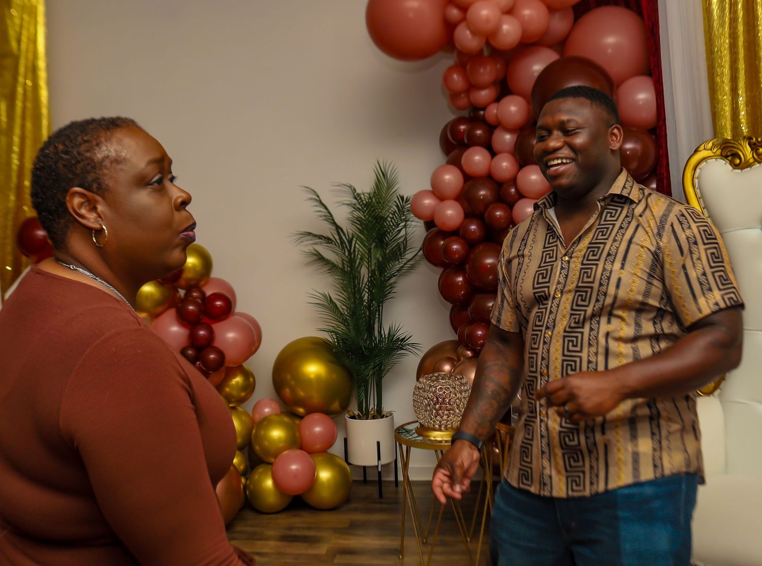 Two people engaged in conversation in front of a colorful balloon arch decoration at a party.