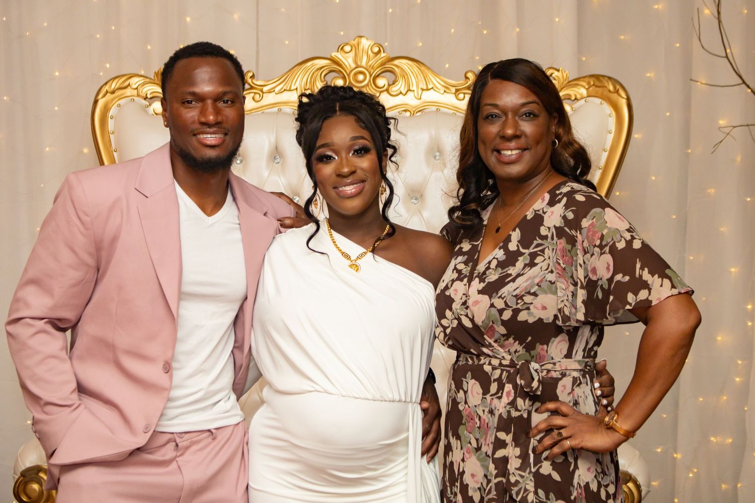 Pregnant woman in white dress poses with two guests at baby shower photo backdrop.