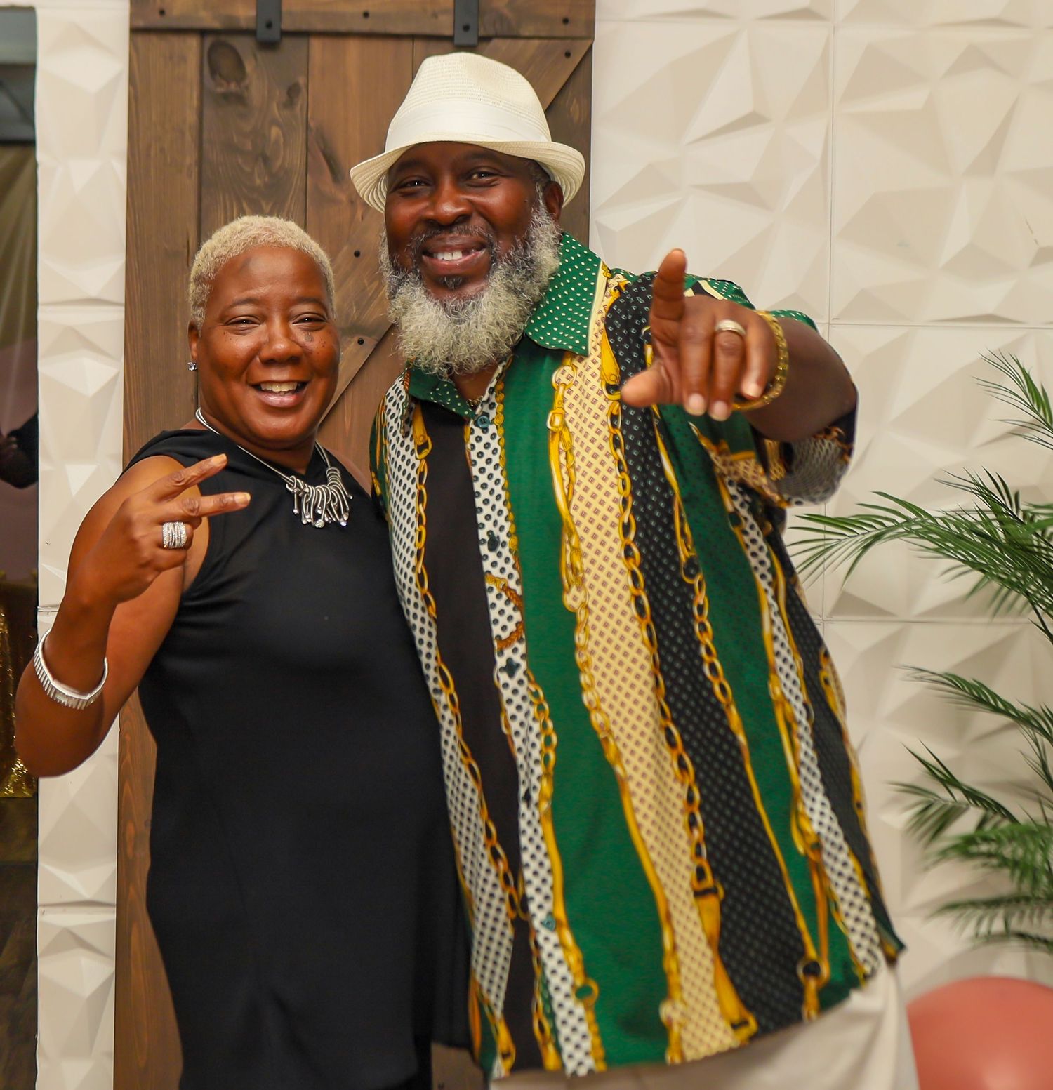 Smiling couple posing together at a party, man wearing colorful striped shirt and white fedora hat.