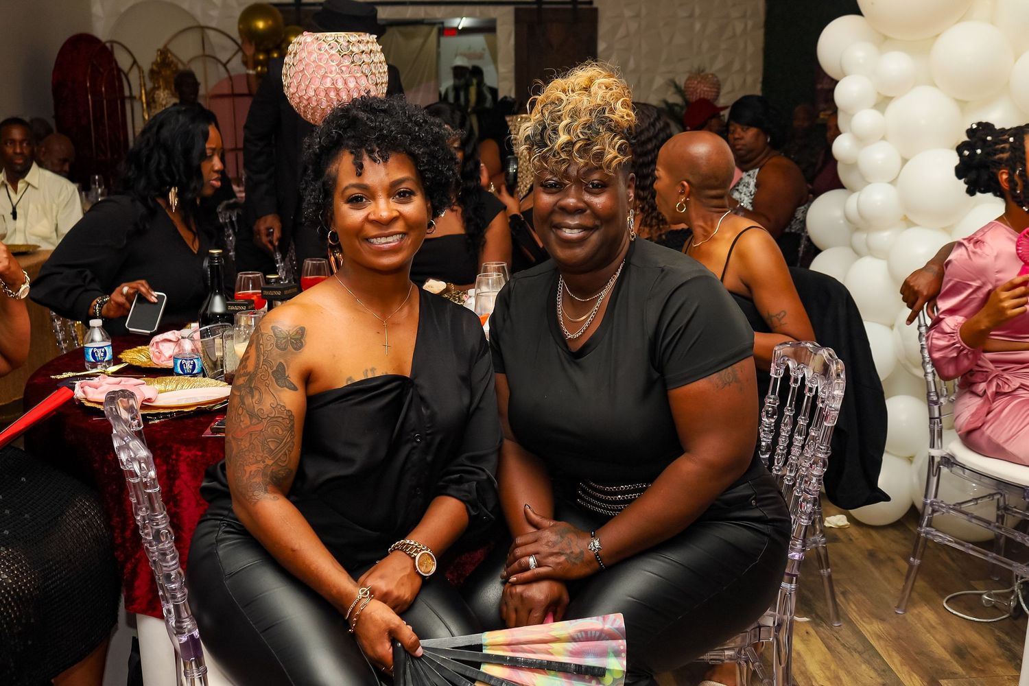 Two women in black outfits smiling together at a festive celebration with balloon decorations.