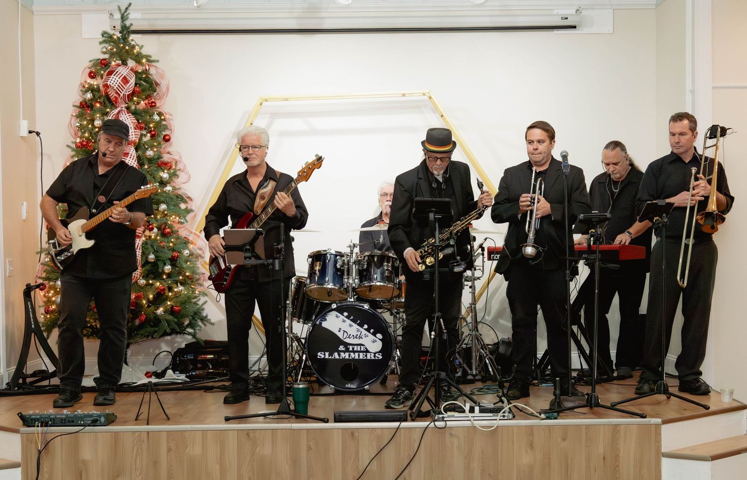 Seven-piece band performing holiday music in front of Christmas tree at indoor venue event.