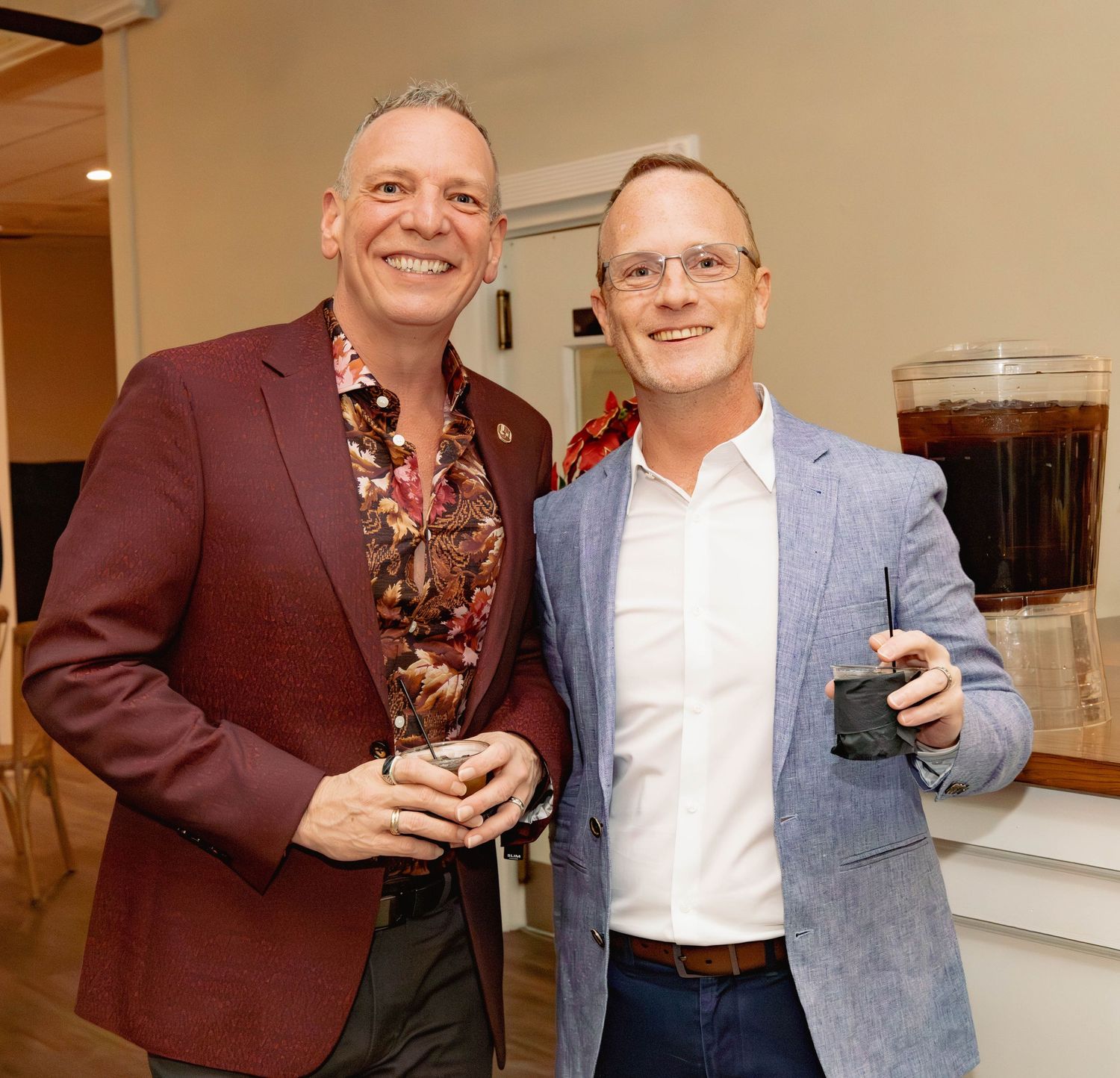 Two men smiling and holding drinks at an indoor holiday party event wearing blazers.
