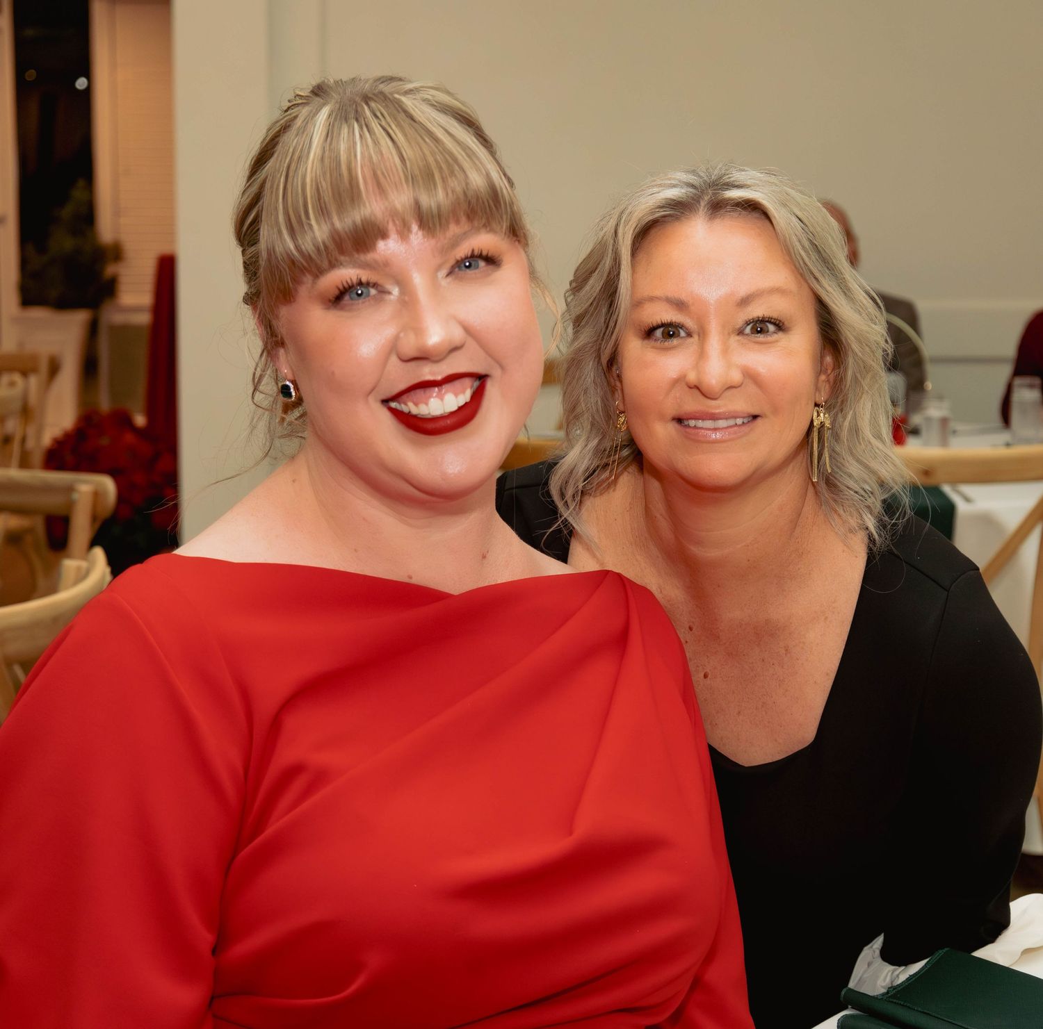 Two women smiling at a festive holiday party, one in red dress and one in black outfit.