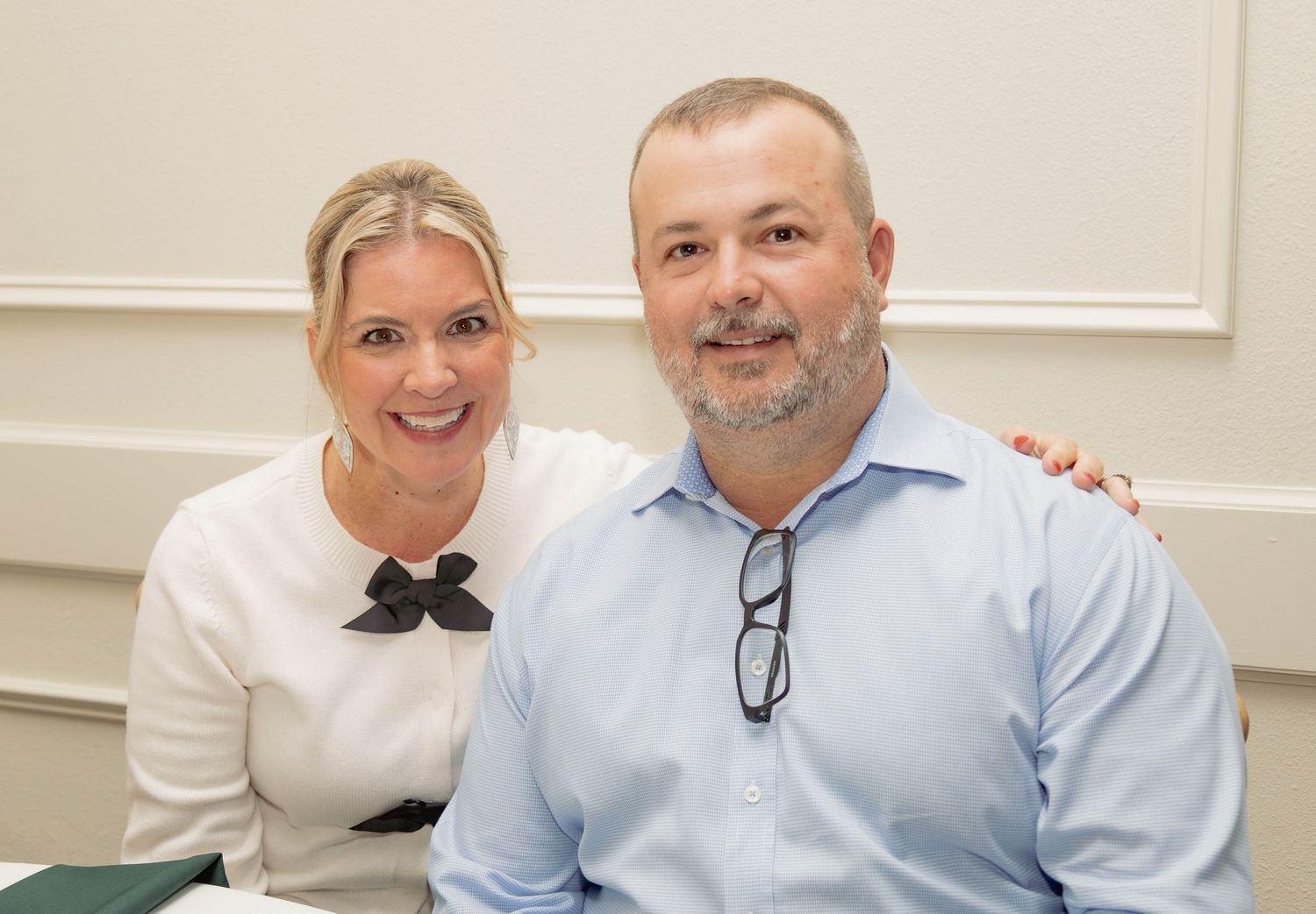 Smiling couple posing together at an elegant event, woman in white blouse and man in blue shirt.