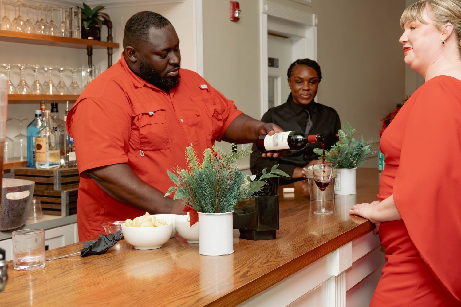 Chef in red uniform pouring wine at bar while staff assists guest at catered event.
