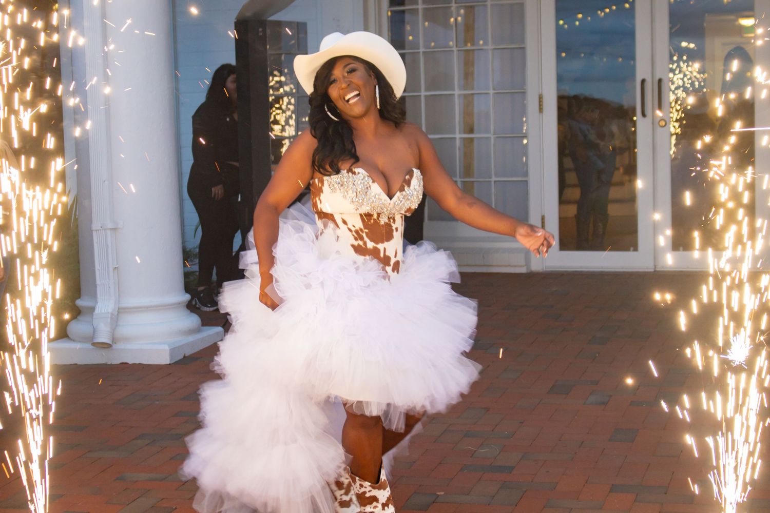 Woman in white tulle dress and cowboy hat celebrating amid sparkling fireworks.