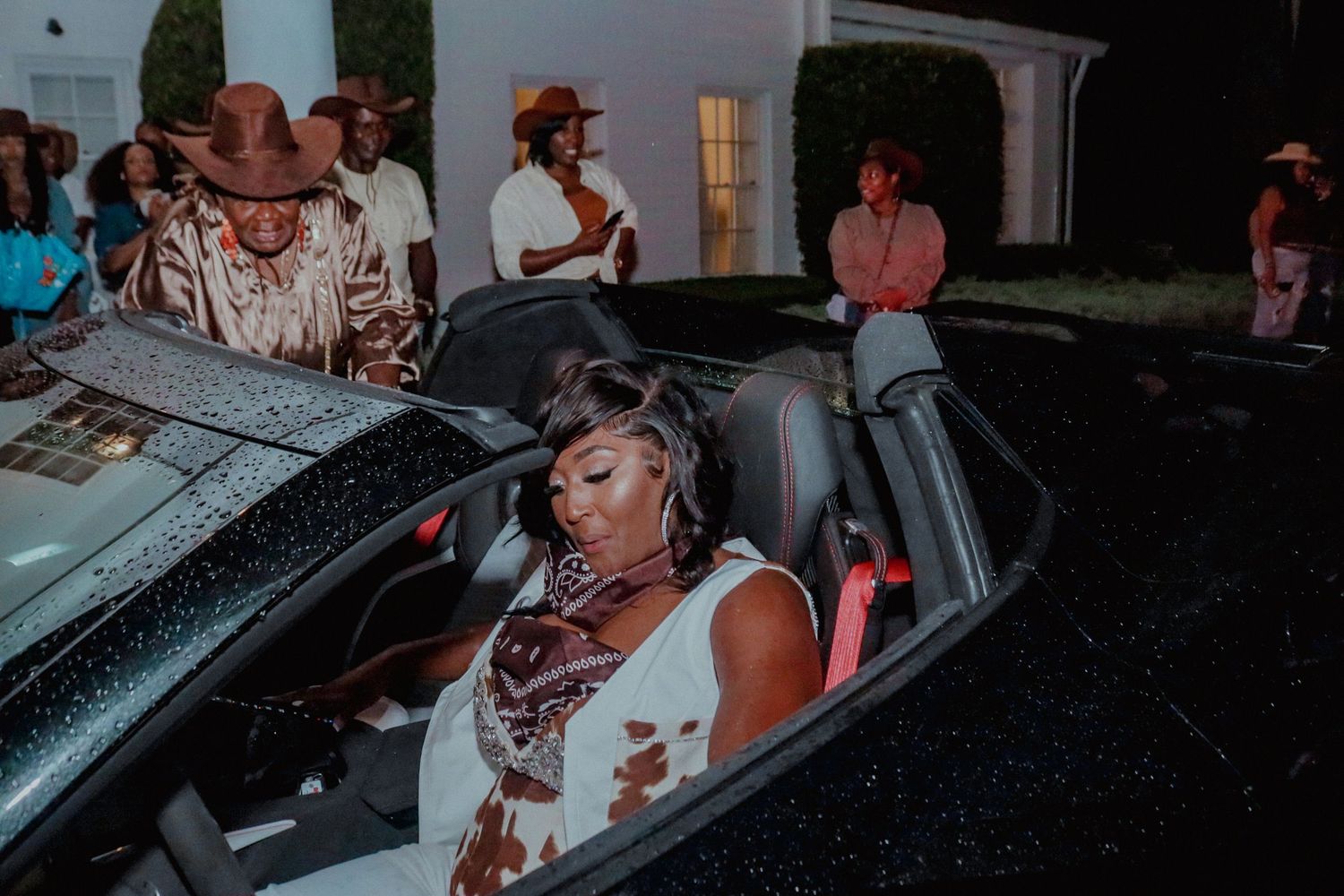 Woman in white dress sitting in a black convertible sports car at an outdoor evening party event.