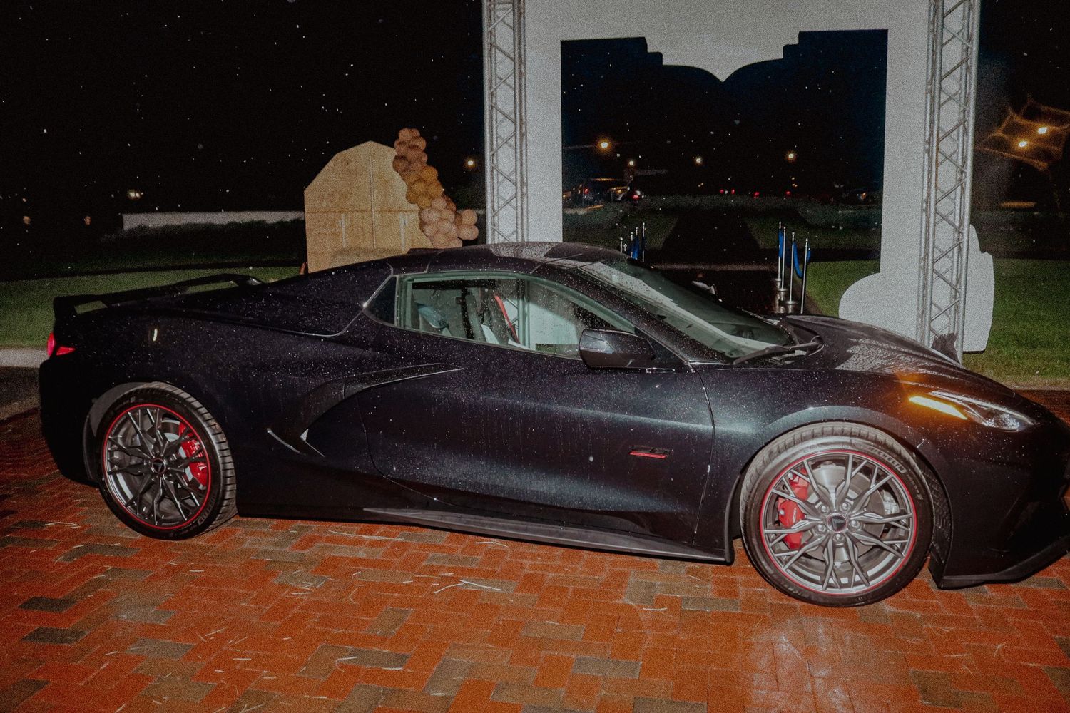Black Chevrolet Corvette convertible sports car parked on brick pavement outside building at night.