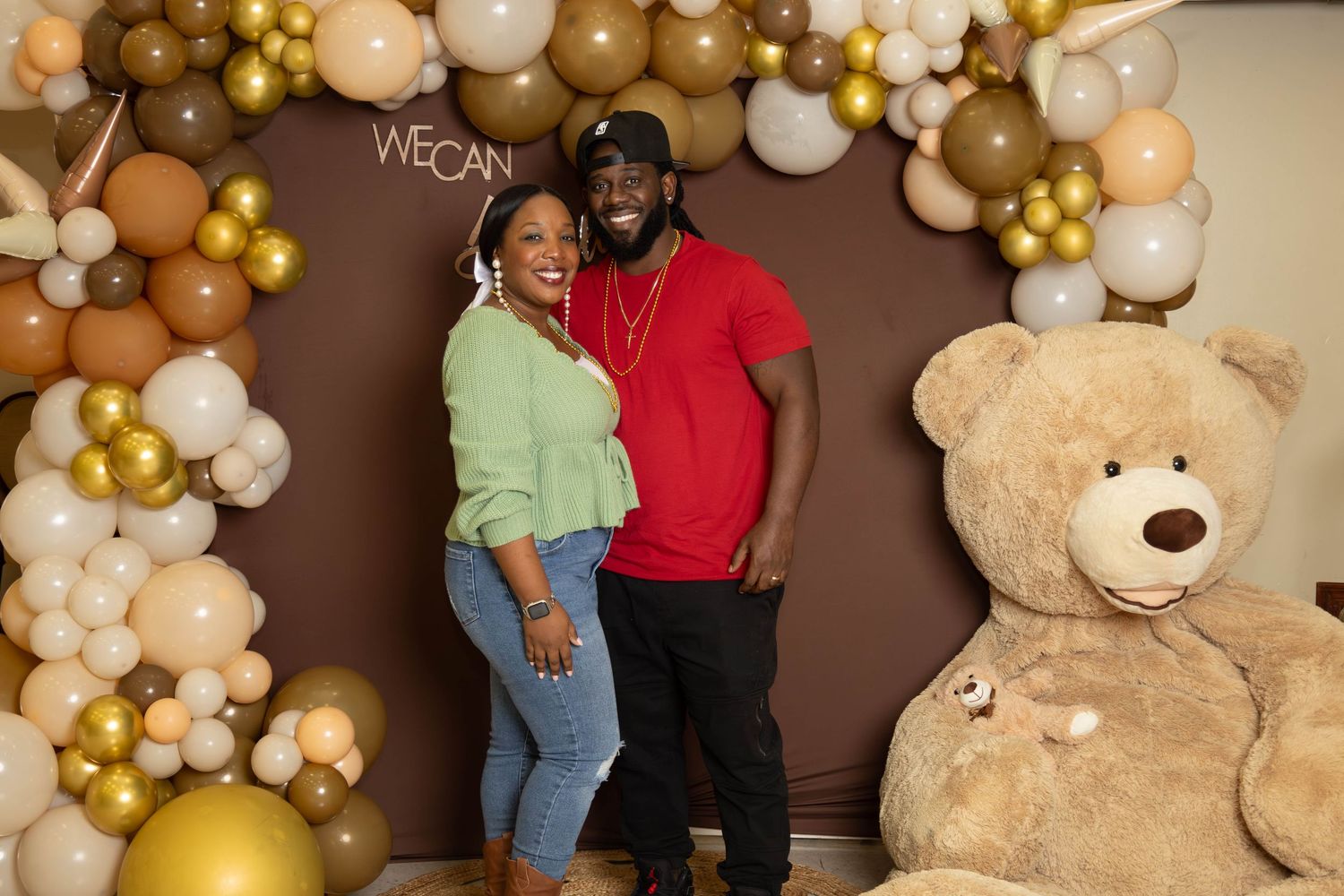 Couple posing in front of a gold and brown balloon arch with a large teddy bear at a baby shower.