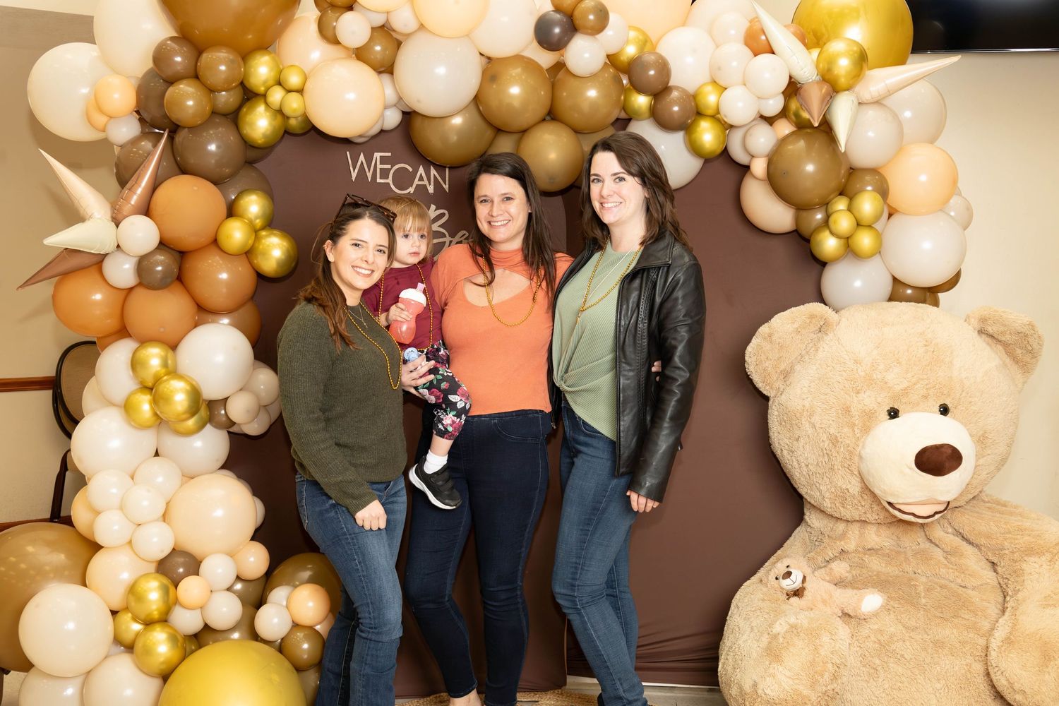 Three women posing with a baby in front of a gold balloon arch and large teddy bear.