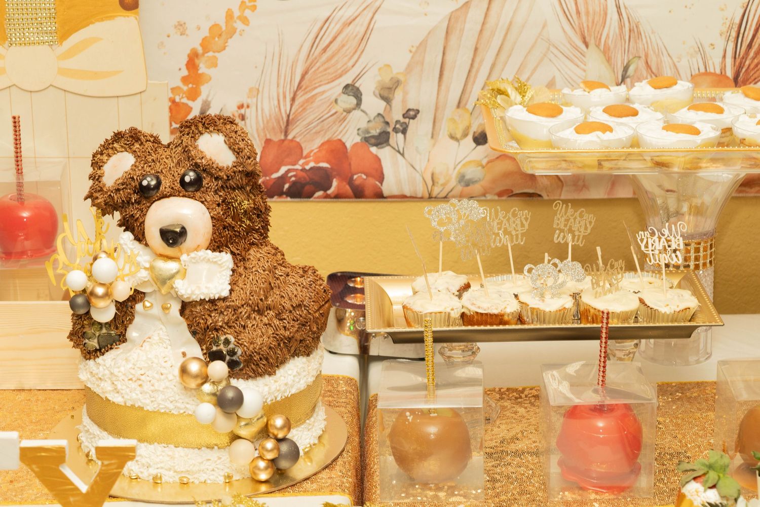 Adorable teddy bear shaped cake with gold accents displayed at a baby shower dessert table.