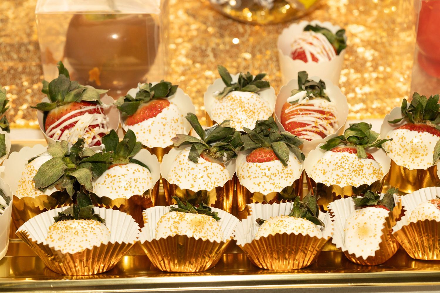 White chocolate dipped strawberries with gold sprinkles in gold cupcake liners on a dessert table.