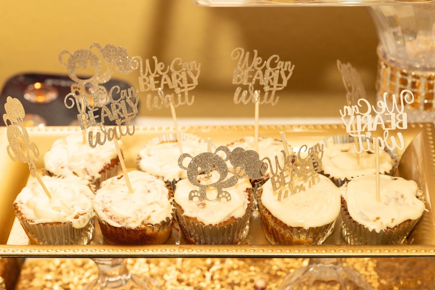 Baby shower cupcakes with bear themed toppers reading We Can Bearly Wait on a gold tray.