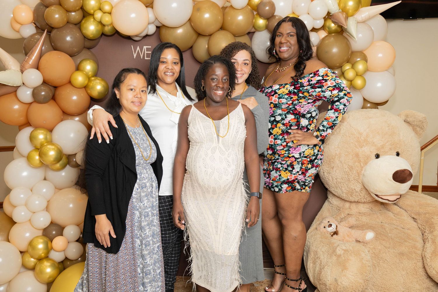 Group of five women posing together in front of a gold balloon arch at a baby shower.