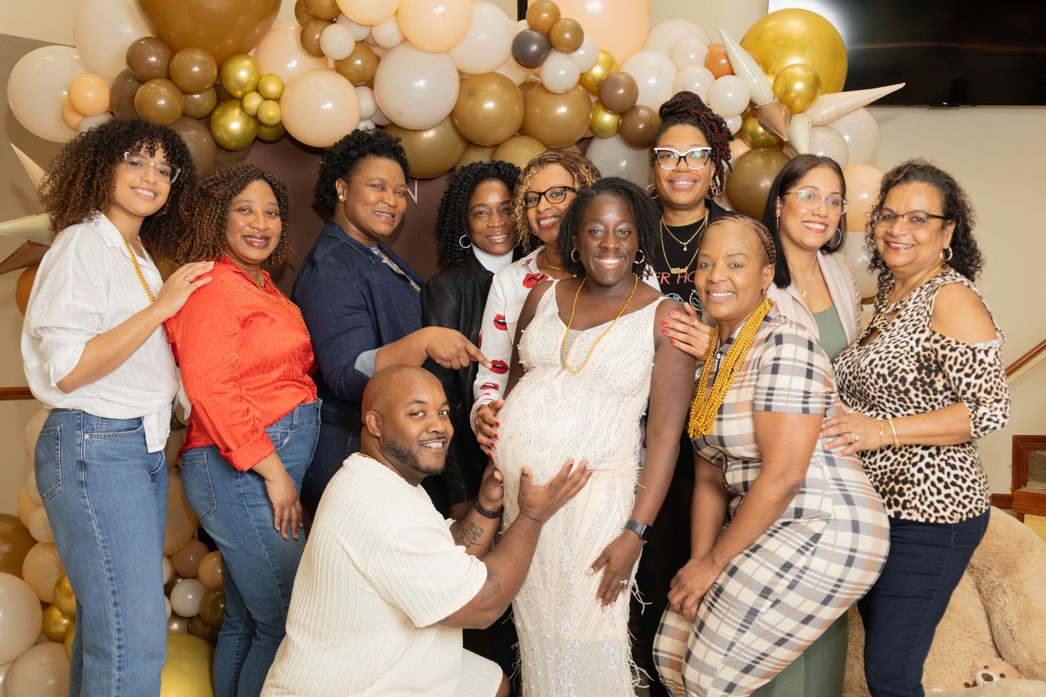A joyful group of people posing together at a baby shower celebration with gold and white balloon decorations.