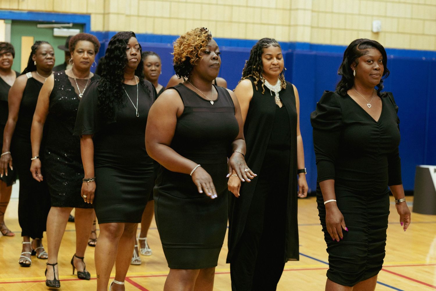 Group of women dressed in black dresses walk together at a formal event in a gymnasium.