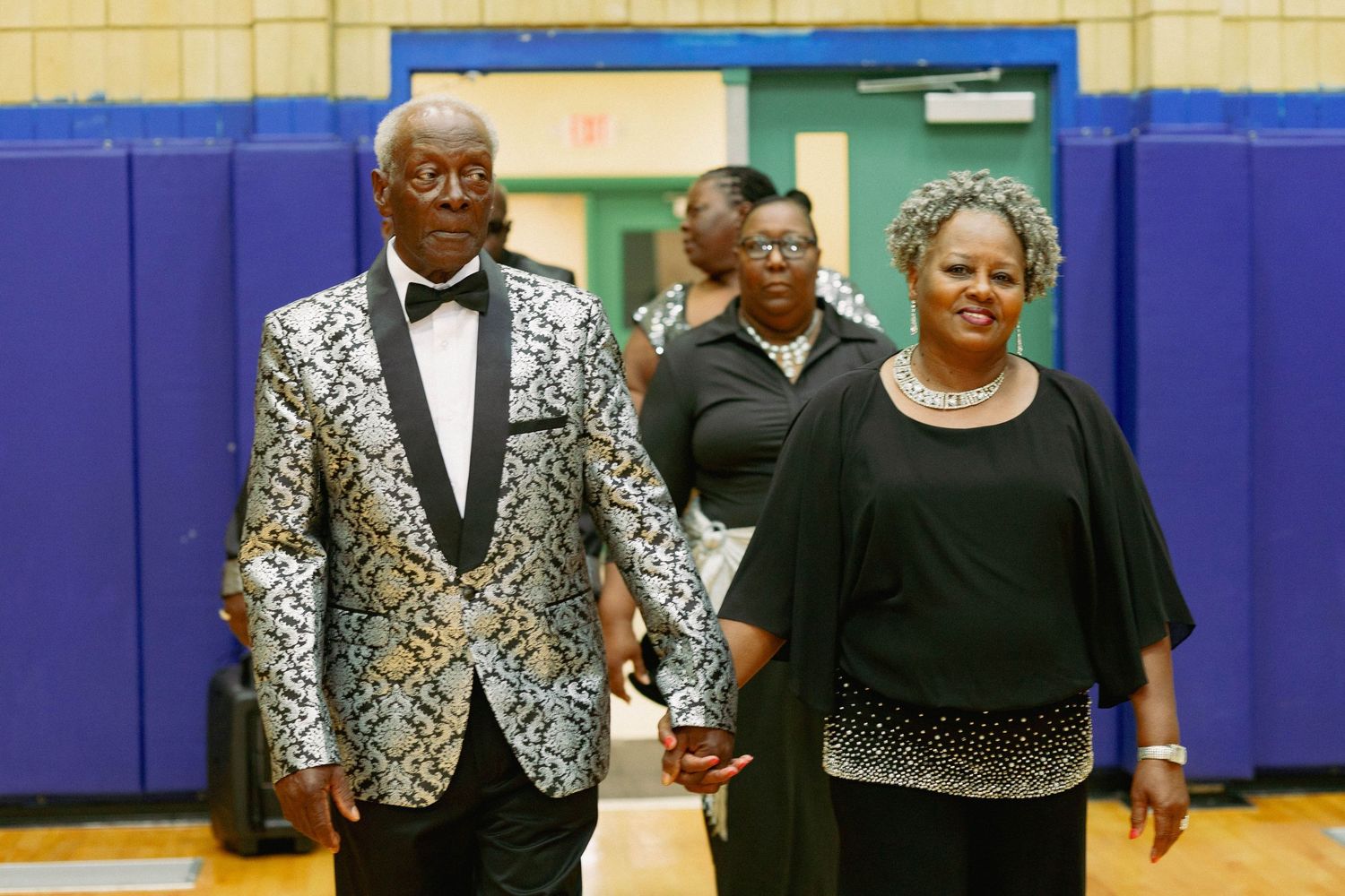 Elegantly dressed couple holding hands walking into a formal celebration event.