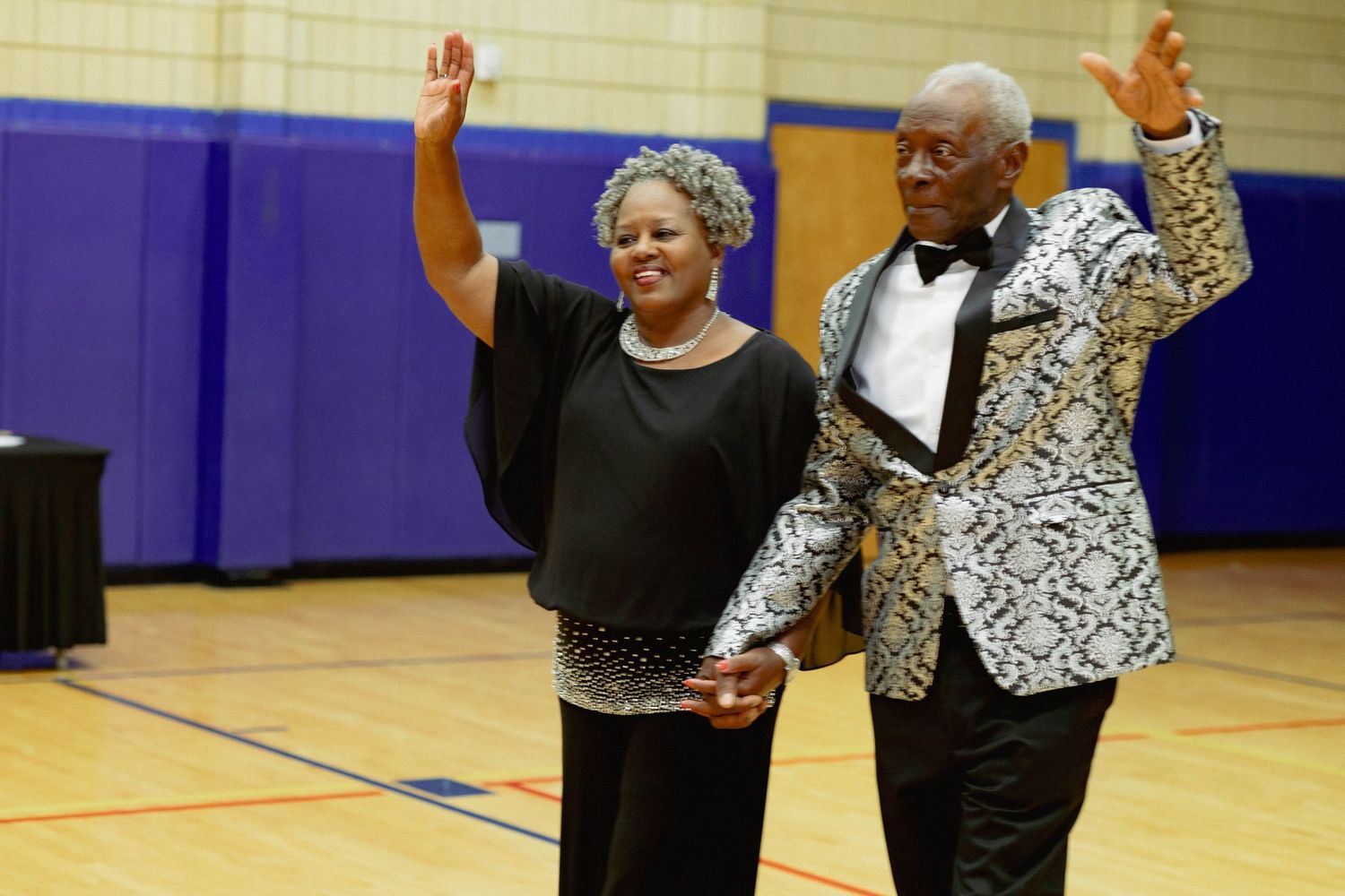 Joyful couple in formal attire waving to guests at an 80th birthday celebration.