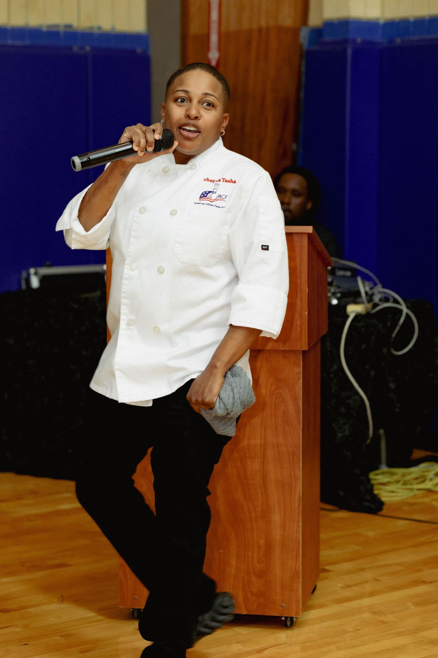 Female chef in white uniform speaks into microphone at a special event.