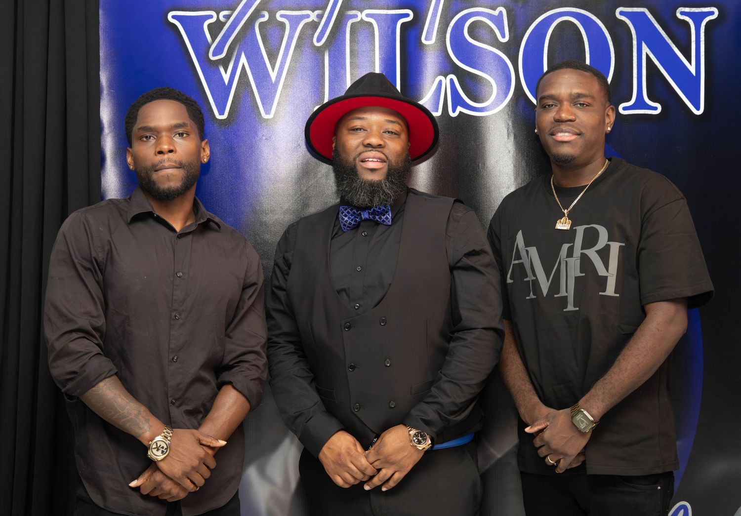 Three young men posing in front of a Wilson sign at a formal event or celebration.