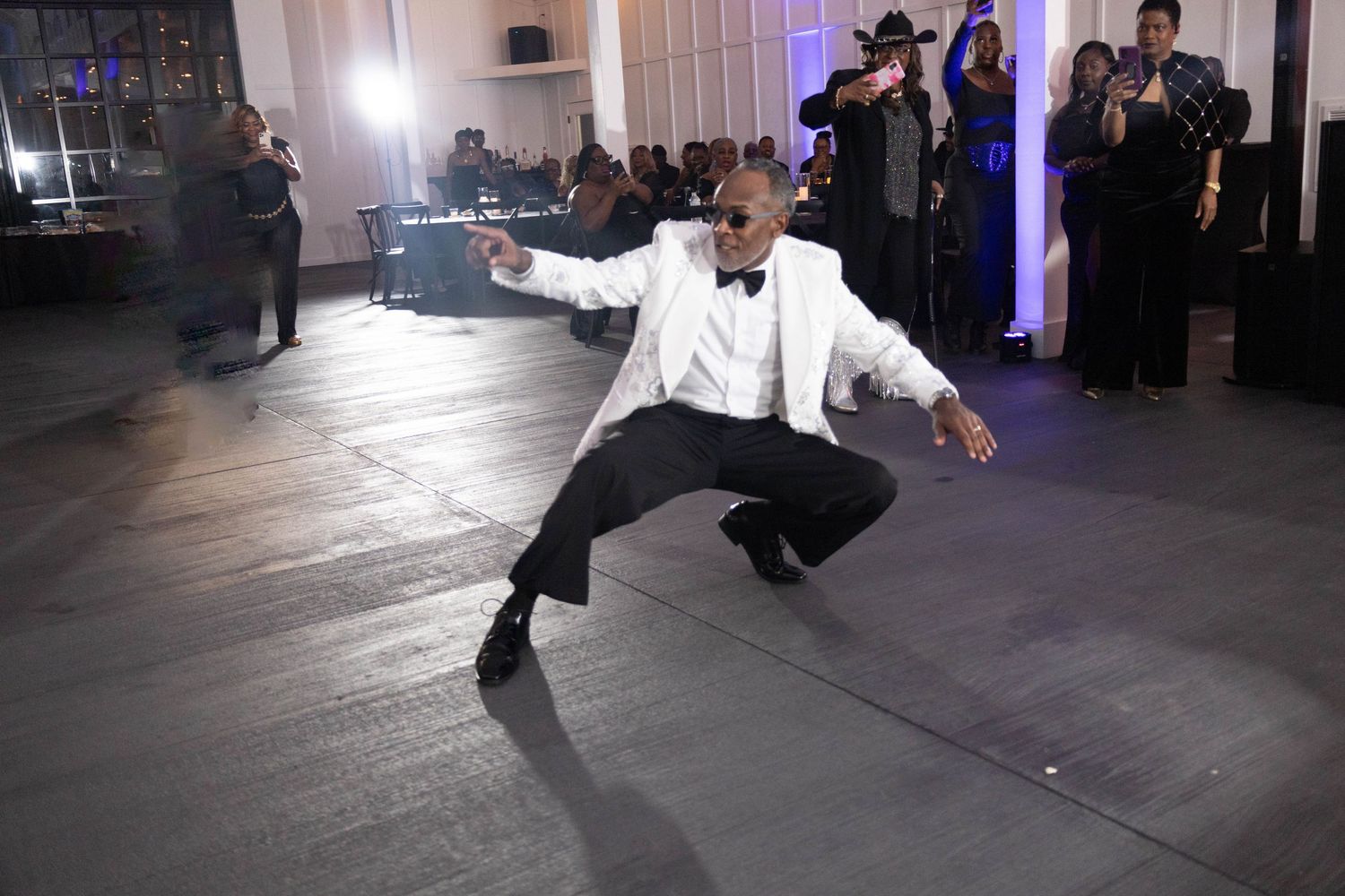 Man in white tuxedo jacket performing an energetic dance move at a formal gala event.