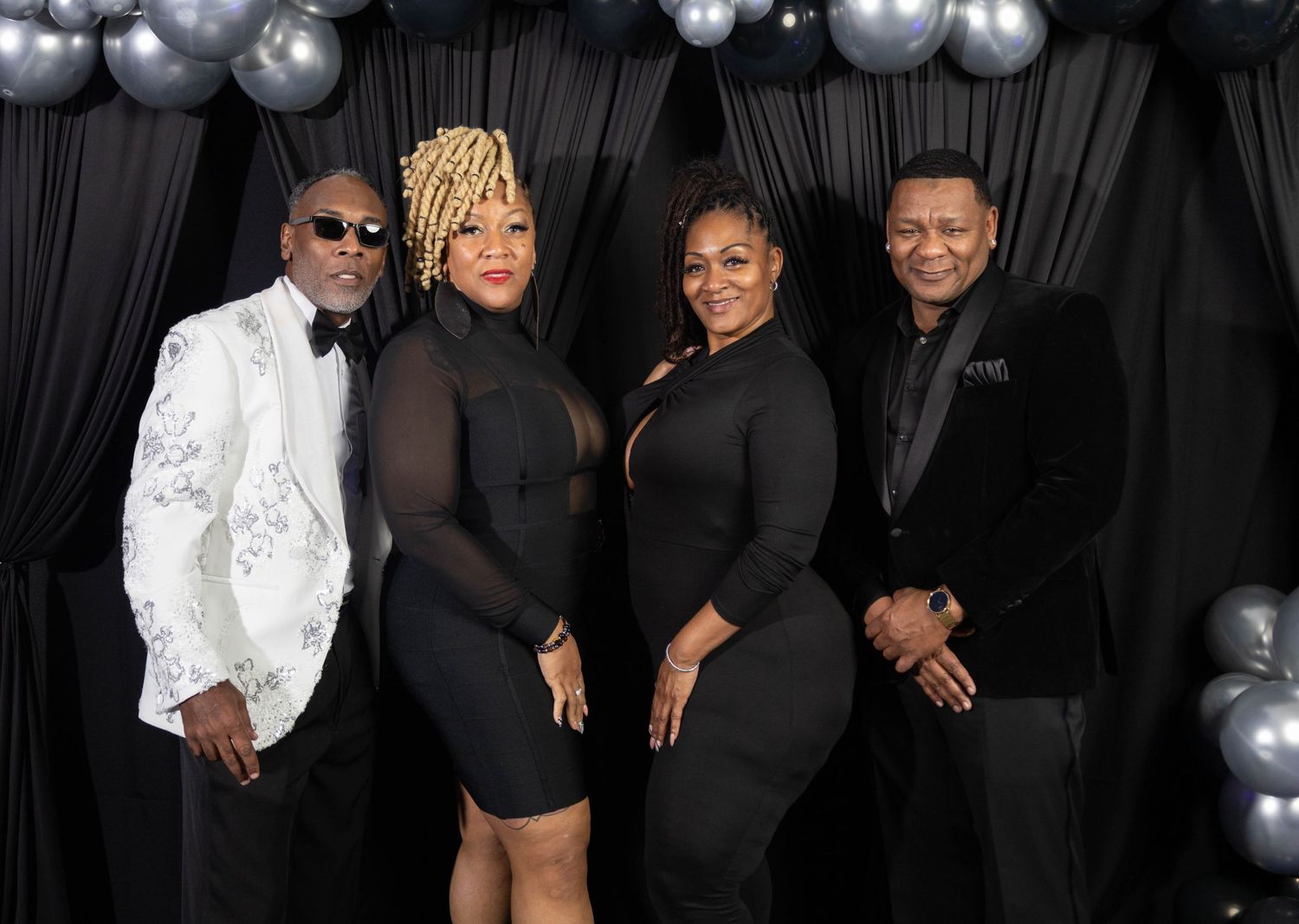 Four elegantly dressed people posing together at a formal event with black and silver balloon decorations.