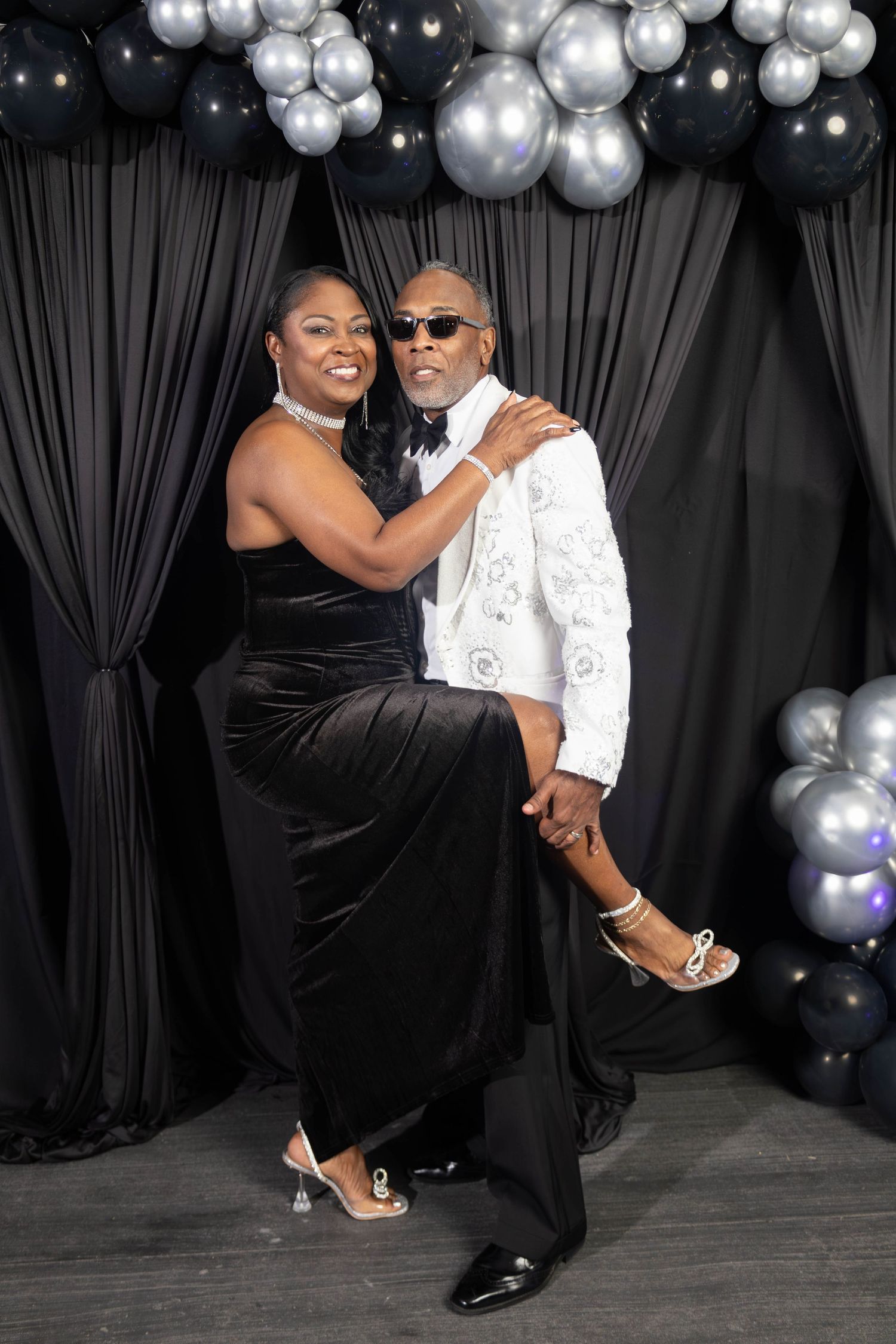 A couple posing playfully at a formal event with black and silver balloon decorations in the background.