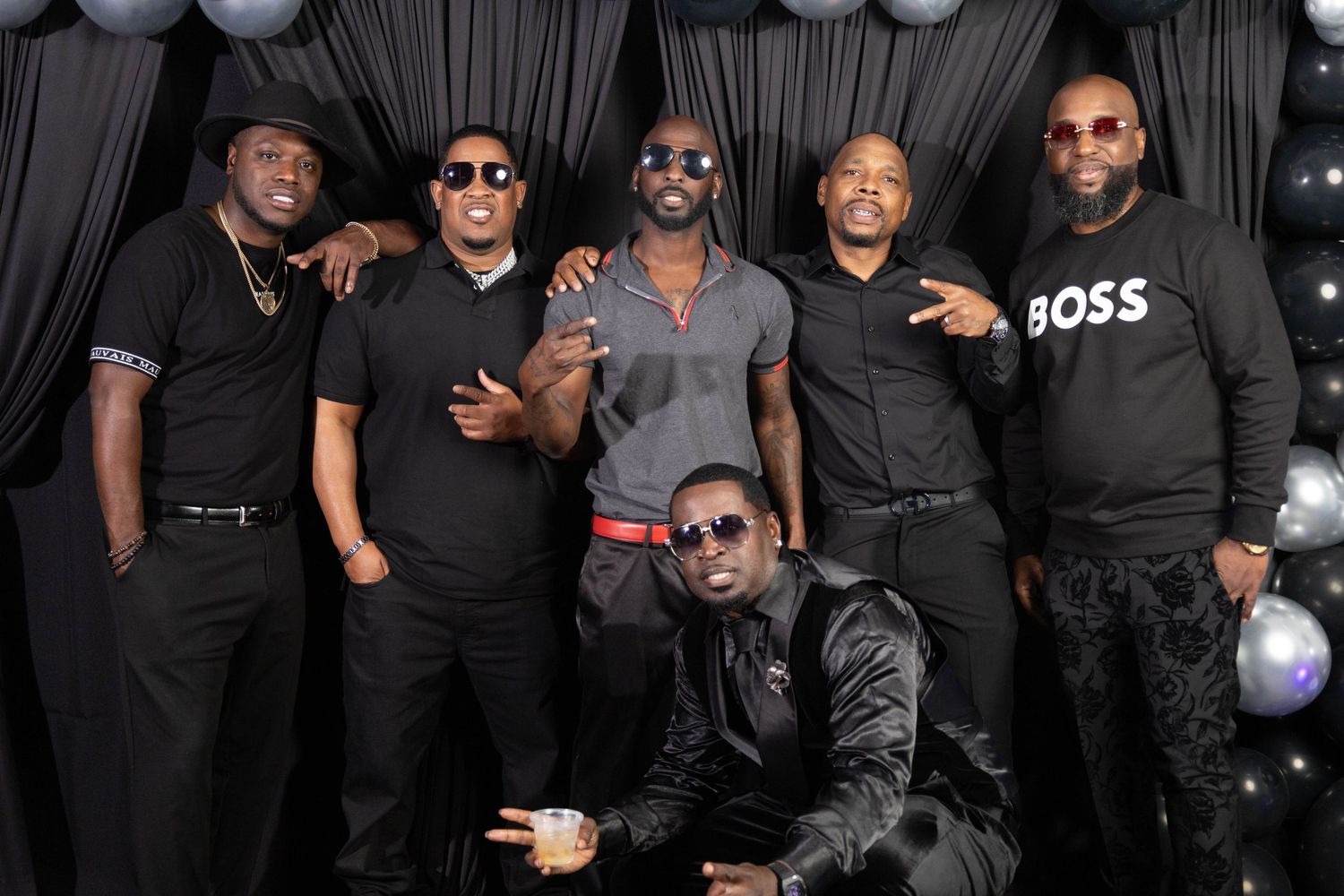Group of six men dressed in black posing together at a formal event with draped backdrop.