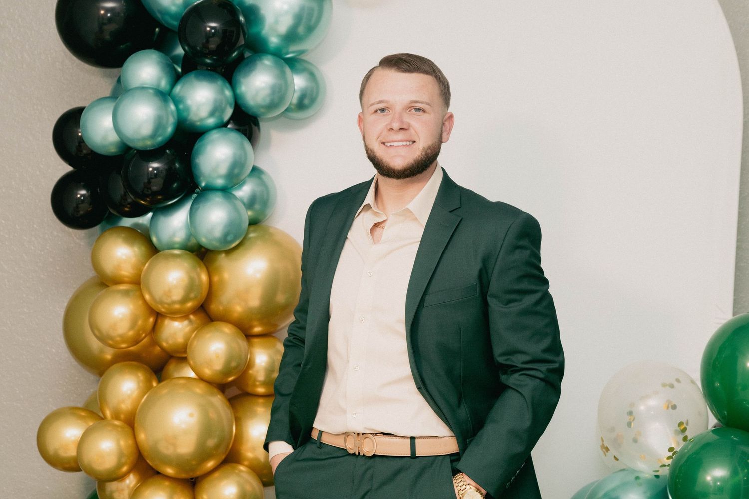 Young man in a green suit posing beside gold and green balloon decorations at a celebration.