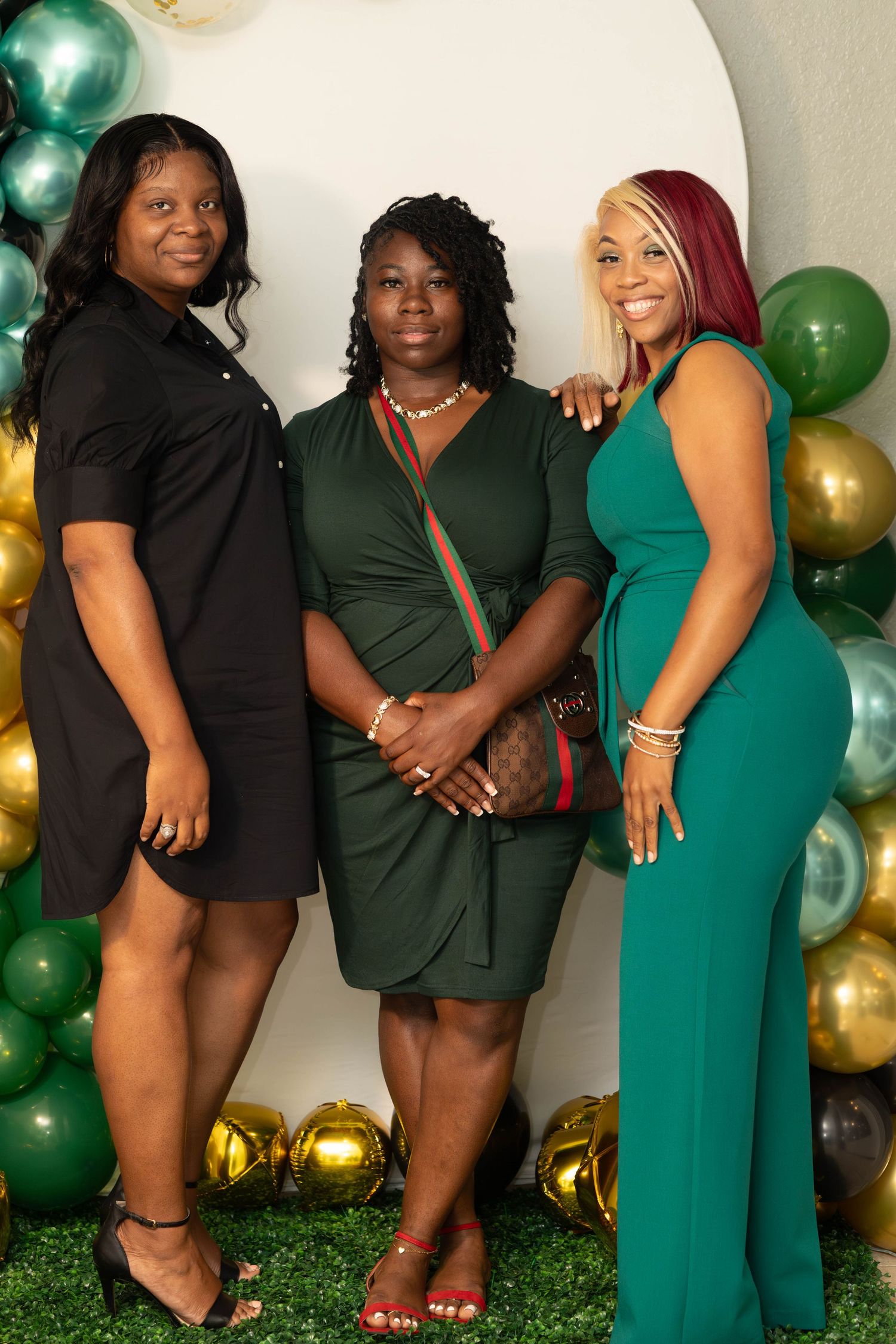 Three women posing together at a green and gold balloon backdrop party event.
