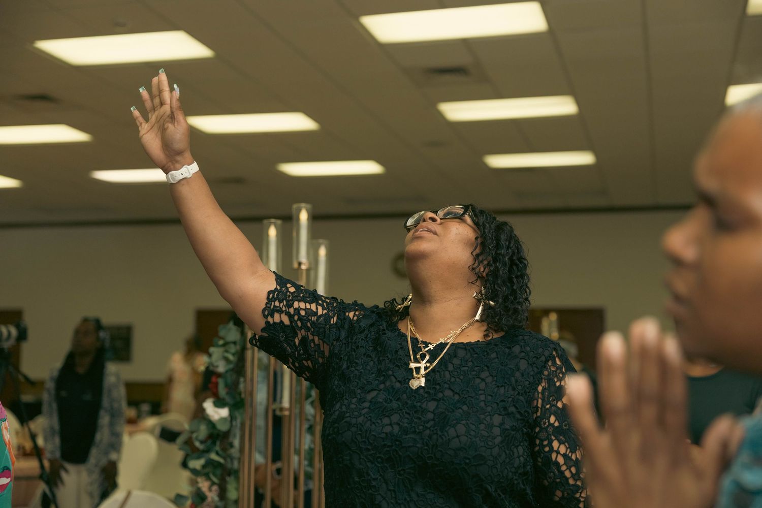 Woman raising hand in worship at a religious gathering with others praying nearby.